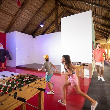 Impressive Punta Cana Hotel Exterior photo The photo shows an indoor recreational space with a thatched roof. In the foreground, there is a foosball table with figures attached to it, where two children are playing or running towards it. A man and a woman, likely parents, are walking together