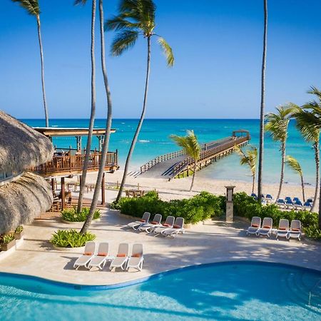 Impressive Punta Cana Hotel Exterior photo The photo depicts a serene tropical scene featuring a swimming pool in the foreground, surrounded by lounge chairs. In the background, there is a beautiful beach with clear turquoise waters and a wooden pier extending out into the ocean. Palm trees f