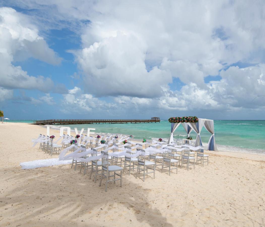 Impressive Punta Cana Hotel Exterior photo The photo shows a beach setting that appears to be prepared for a wedding ceremony. There are rows of white chairs arranged on the sand facing a decorated archway. The arch is draped in white fabric and adorned with floral elements. In the background