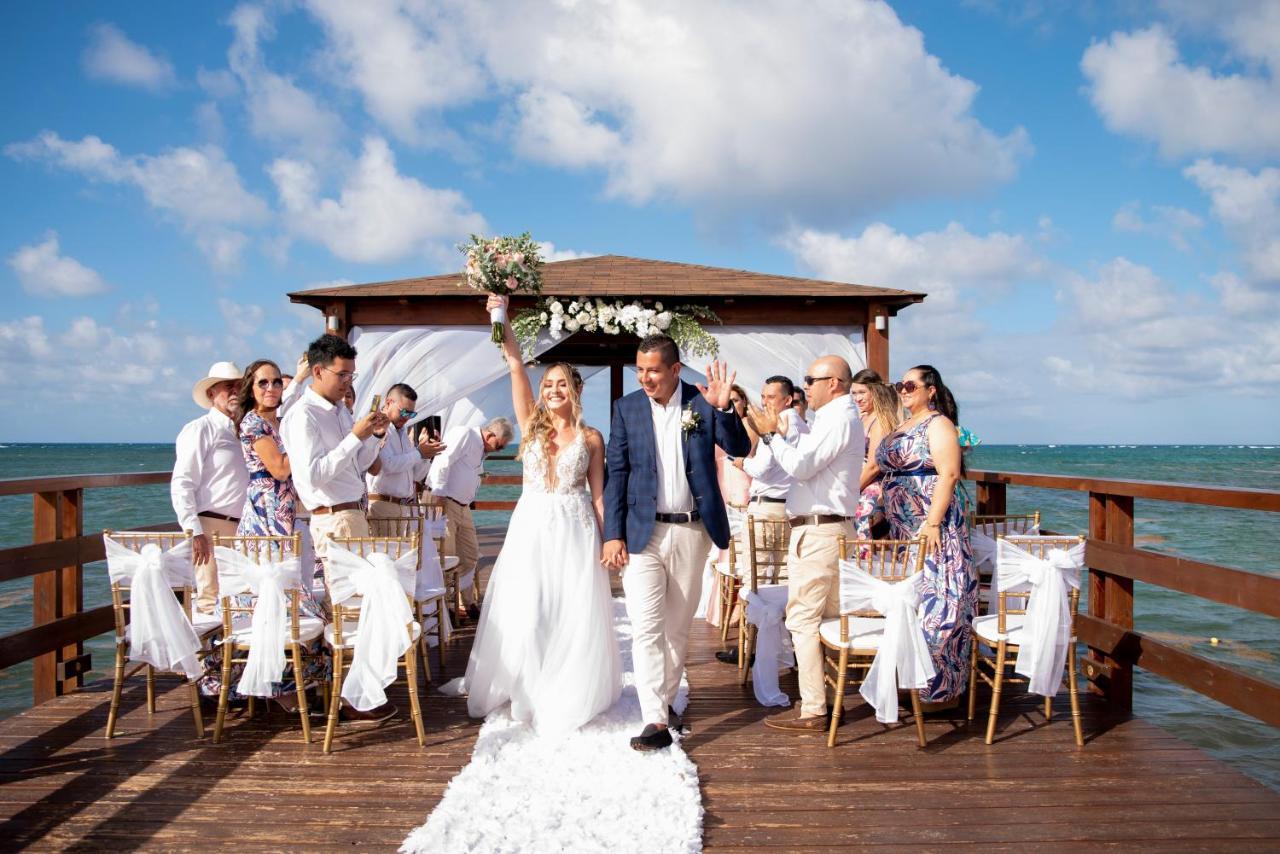 Impressive Punta Cana Hotel Exterior photo The photo shows a wedding ceremony taking place on a wooden pier over water. A bride and groom are at the center, with the bride holding her bouquet up in celebration. They are surrounded by a group of guests, all dressed in formal attire, some clapp