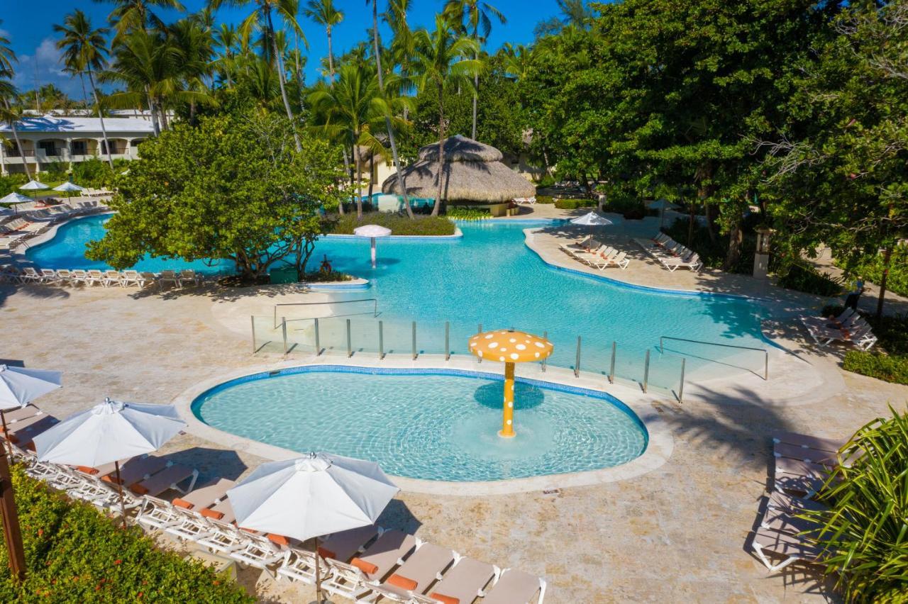 Impressive Punta Cana Hotel Exterior photo The photo shows a vibrant outdoor pool area surrounded by lush greenery. There are two distinct pool sections visible: a larger main pool with a clear blue water and a smaller, circular pool that features a playful mushroom-shaped fountain. Sun loung