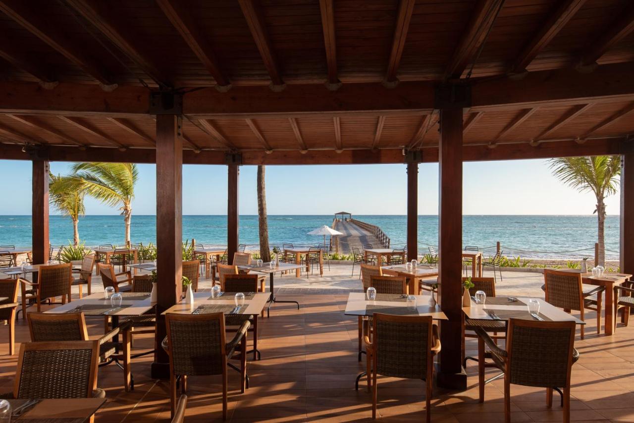 Impressive Punta Cana Hotel Exterior photo The photo shows a beachfront dining area with a wooden structure featuring a thatched roof. There are several tables and chairs arranged on a wooden deck, providing a picturesque view of the ocean. Palm trees can be seen swaying in the breeze, and th