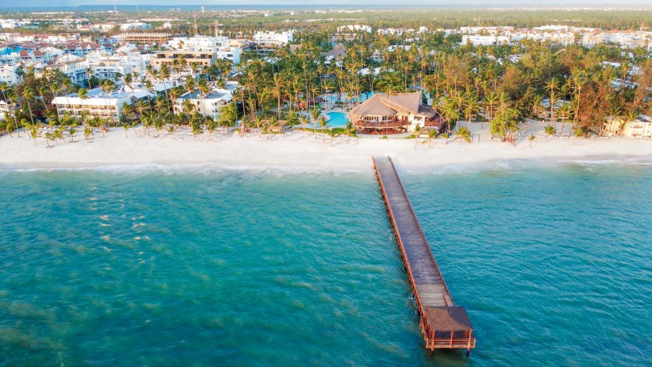 Impressive Punta Cana Hotel Exterior photo The photo shows a picturesque beach scene viewed from above. In the foreground, there is a wooden pier extending over clear turquoise water. The pier leads towards a coastal area with a sandy beach lined by lush palm trees. In the background, several