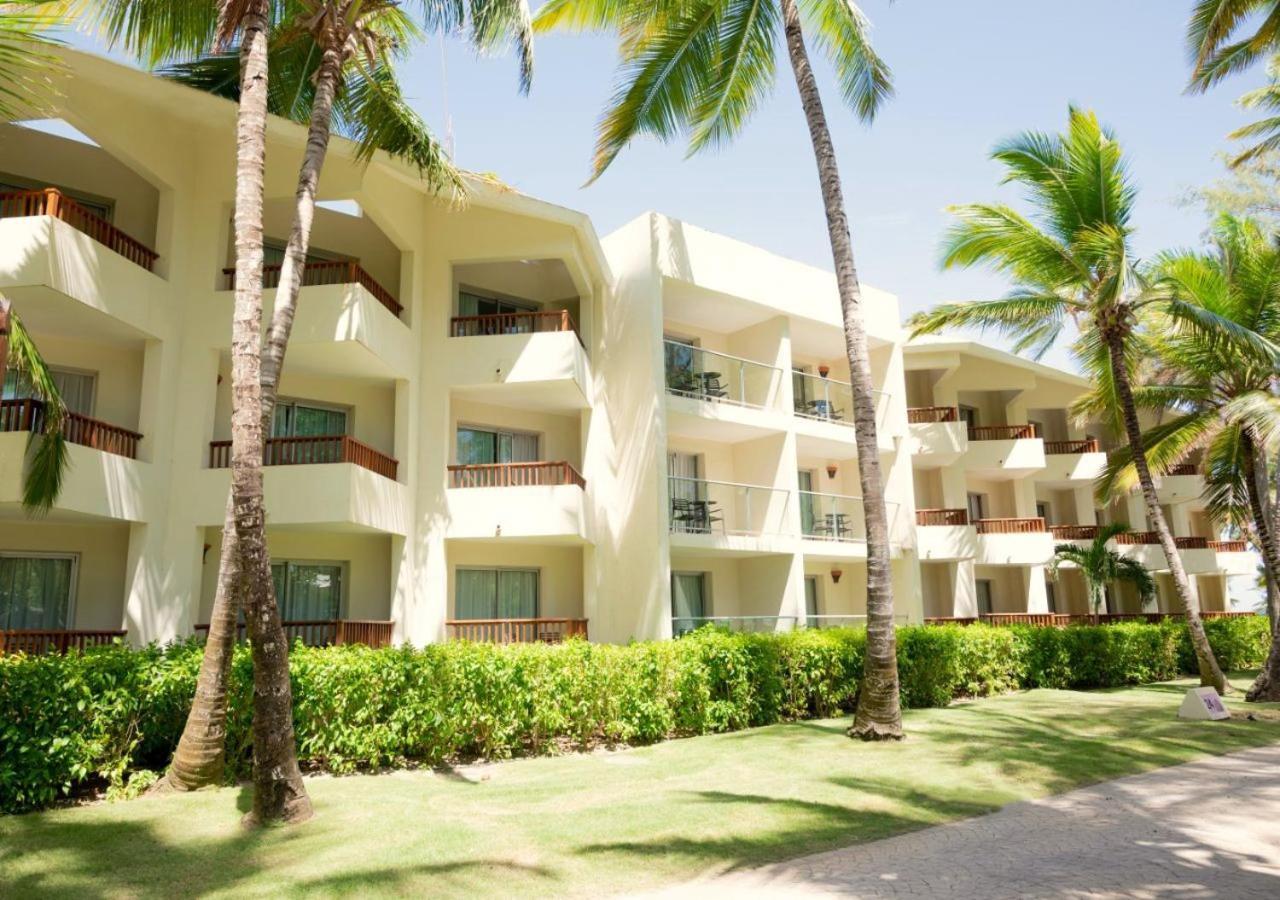 Impressive Punta Cana Hotel Exterior photo The photo shows a beachfront hotel or resort building with multiple levels. The structure is light-colored with balconies that overlook a manicured garden area. Tall palm trees and lush green vegetation surround the building, suggesting a tropical lo