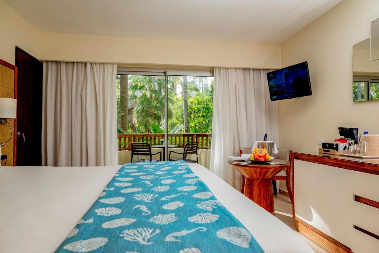 Impressive Punta Cana Hotel Exterior photo The photo shows a cozy hotel room with a large bed adorned with a blue and white decorative blanket featuring shell patterns. In the background, there are large windows with sheer curtains that allow natural light to fill the room, offering a view of