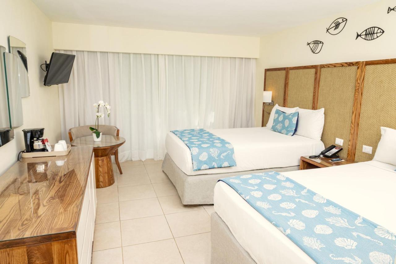 Impressive Punta Cana Hotel Exterior photo The photo shows a hotel room with two beds. Each bed has a light blue bedspread featuring a shell pattern. There is a wooden dresser or side table with a television mounted on the wall. A small round table with a chair is in the corner, next to a win