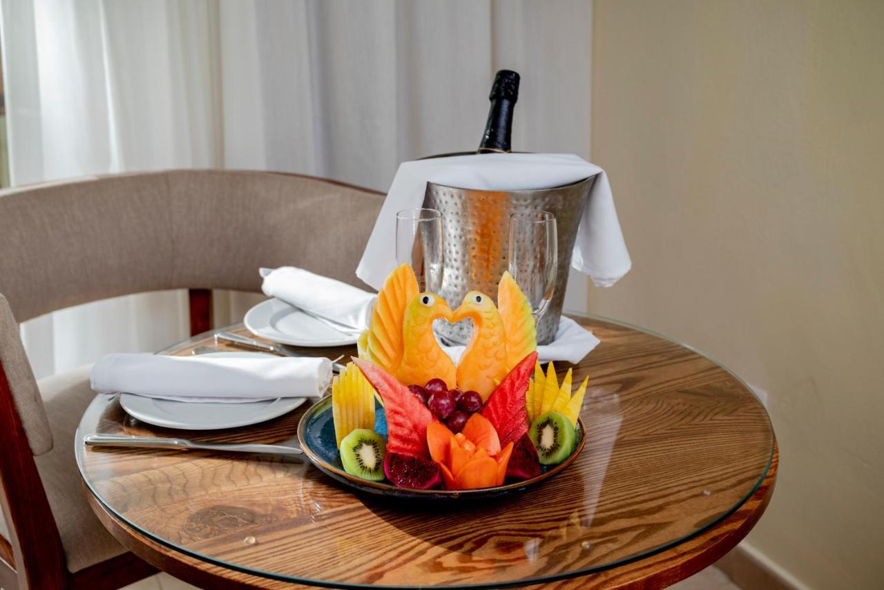 Impressive Punta Cana Hotel Exterior photo The photo shows a cozy table setting. In the center, there is a beautifully arranged platter of fruit art, featuring vibrant pieces of sliced fruits such as mango, kiwi, and various berries, artistically crafted to resemble birds. Next to the fruit a