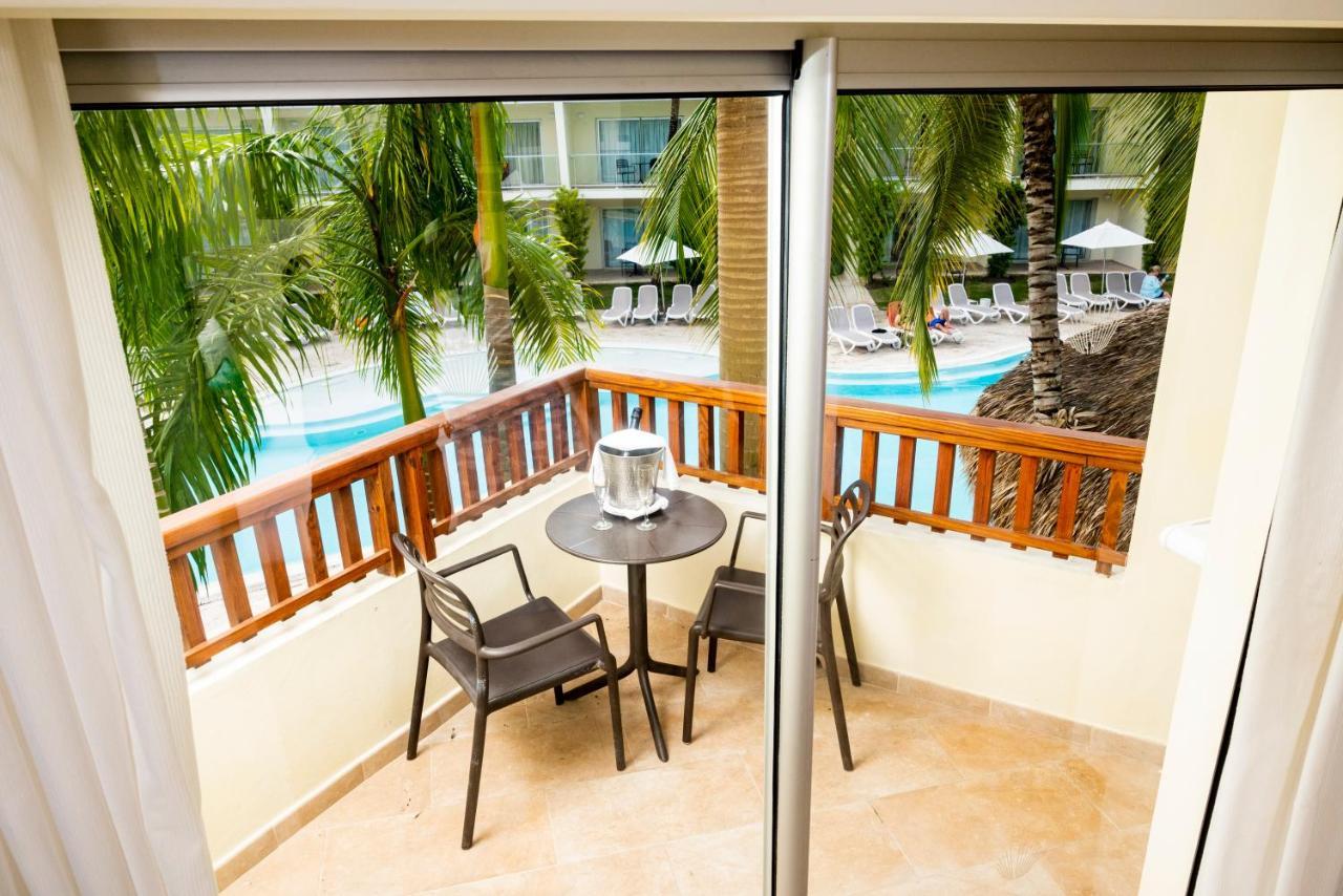 Impressive Punta Cana Hotel Exterior photo The photo shows a small balcony with a table and two chairs. The view from the balcony includes lush green palm trees, and in the background, there is a swimming pool area with lounge chairs. The setting appears to be tropical and relaxing, ideal for