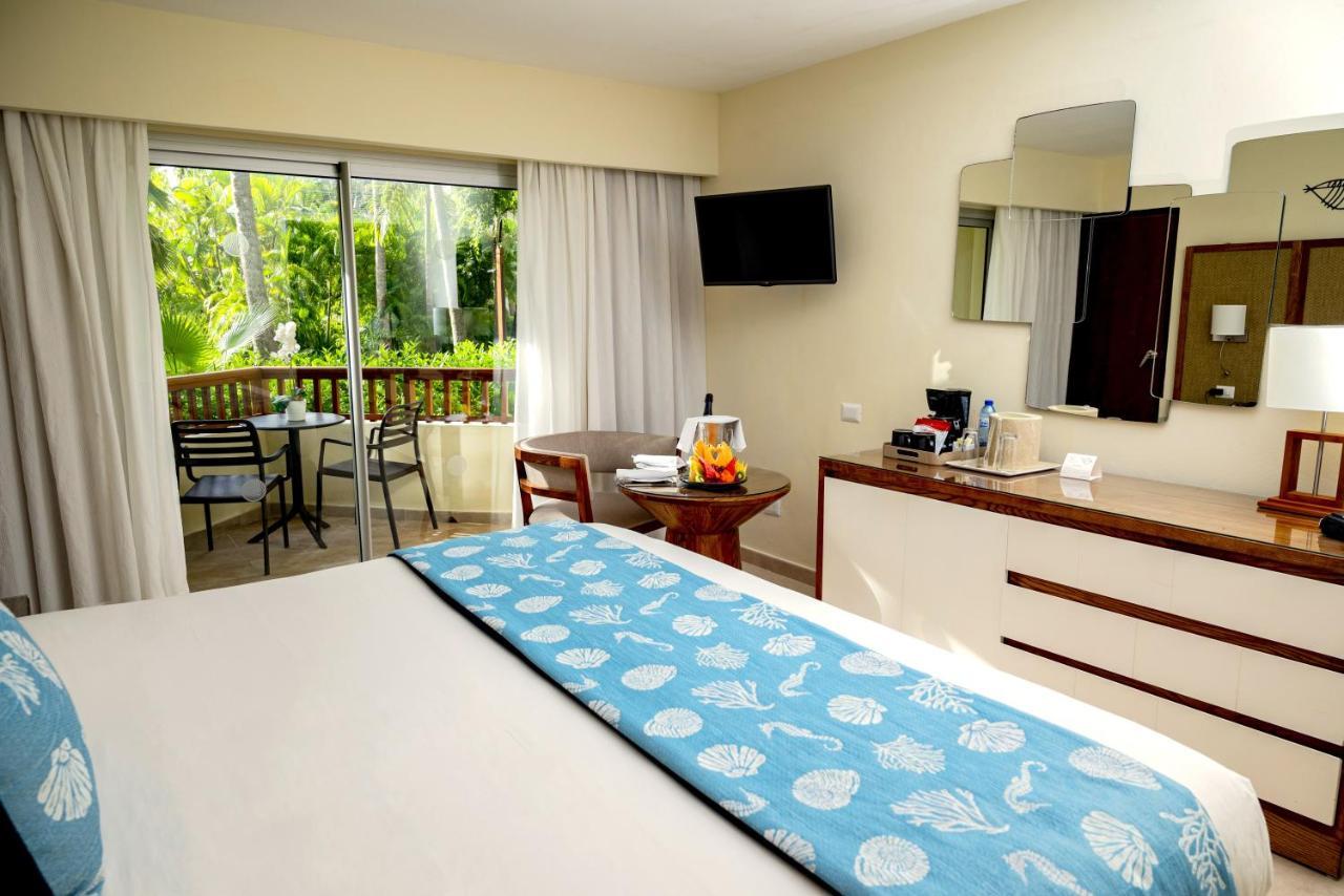 Impressive Punta Cana Hotel Exterior photo The photo shows an inviting hotel room with a bright and airy atmosphere. The room features a large bed with a light-colored headboard and a decorative blue bed runner adorned with shell patterns. There is a wooden dresser with a flat-screen TV mount