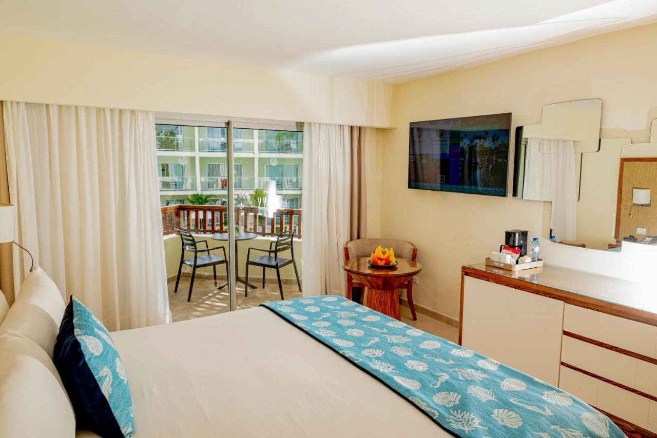 Impressive Punta Cana Hotel Exterior photo The photo shows a hotel room or a well-furnished guest room. It features a comfortable bed with light-colored bedding and a decorative throw in a blue pattern. There is a flat-screen TV mounted on the wall opposite the bed. To the side, there's a sma