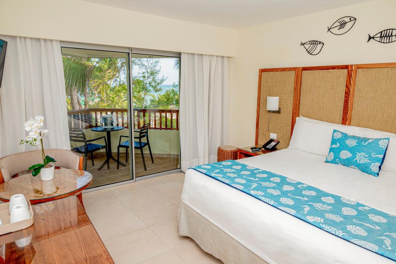 Impressive Punta Cana Hotel Exterior photo The photo shows a cozy hotel room featuring a large bed with a decorative blue and white bed runner. There is a wooden table next to the bed, which holds a small vase with a flower. The room has large sliding glass doors leading to a balcony with out