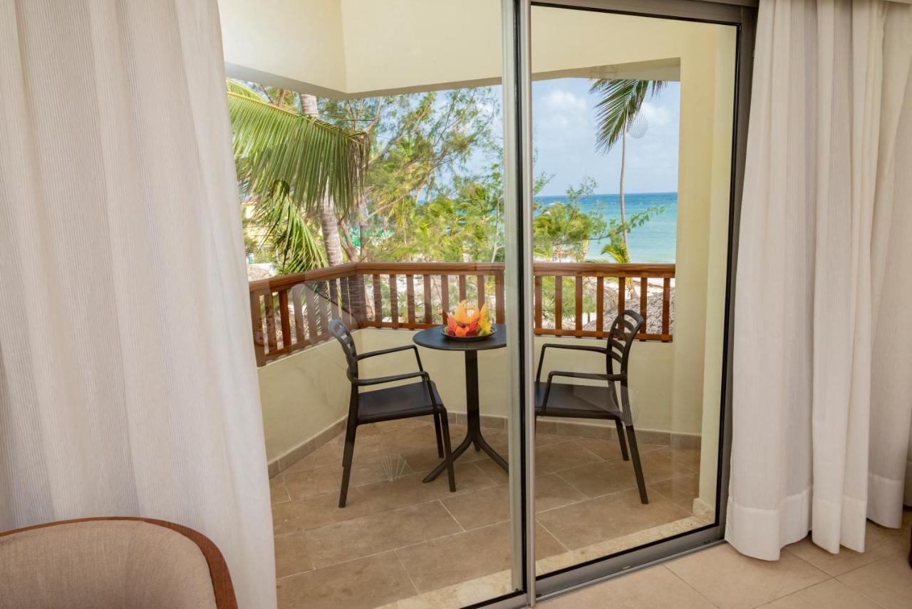 Impressive Punta Cana Hotel Exterior photo The photo shows a balcony with a beautiful view of the ocean. Through glass doors, you can see a small round table with two chairs on the balcony. The setting is bright and inviting, with tropical palm trees visible in the background. The decor appea