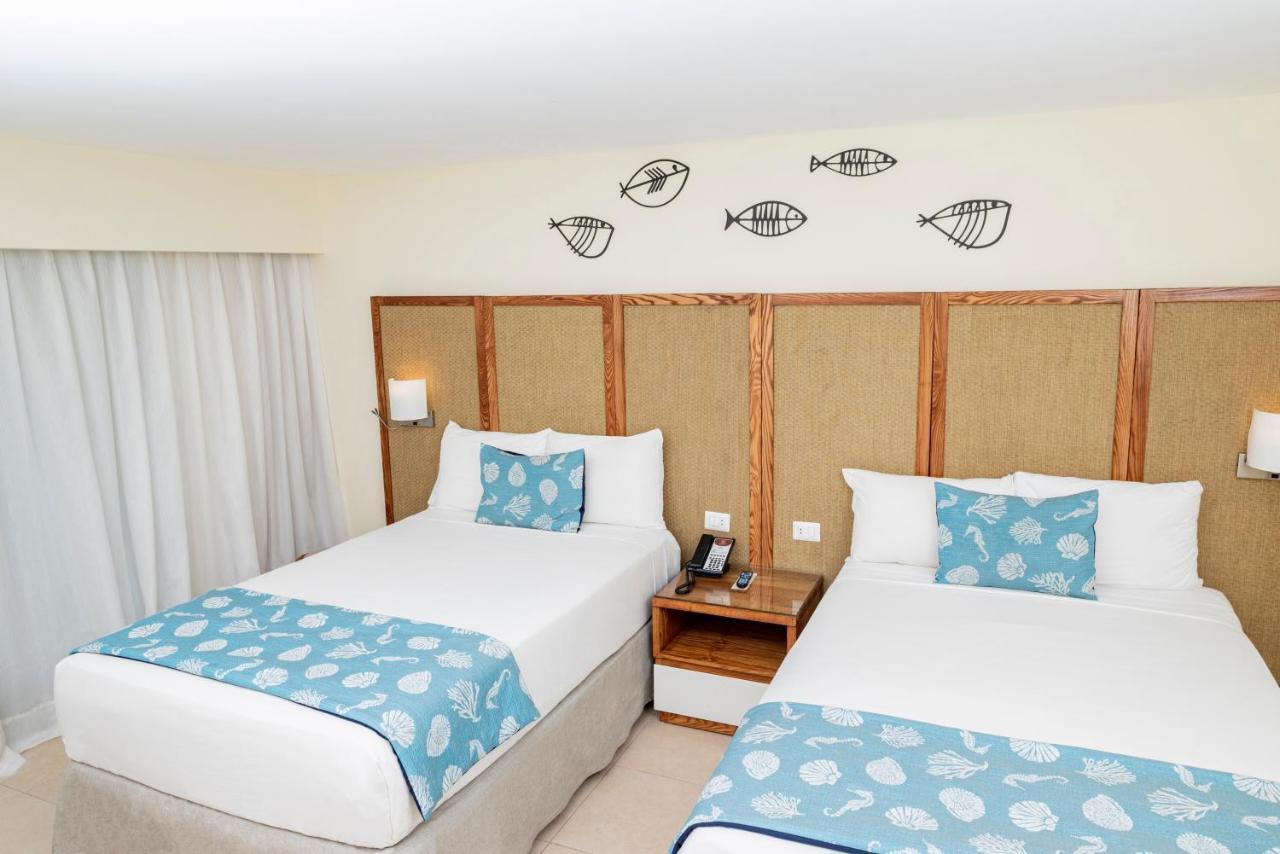 Impressive Punta Cana Hotel Exterior photo The photo shows a hotel room featuring two beds with light-colored bedding. Each bed has a blue decorative pillow with a shell pattern. The walls are adorned with a natural texture, and above the beds, there are decorative fish illustrations. A small