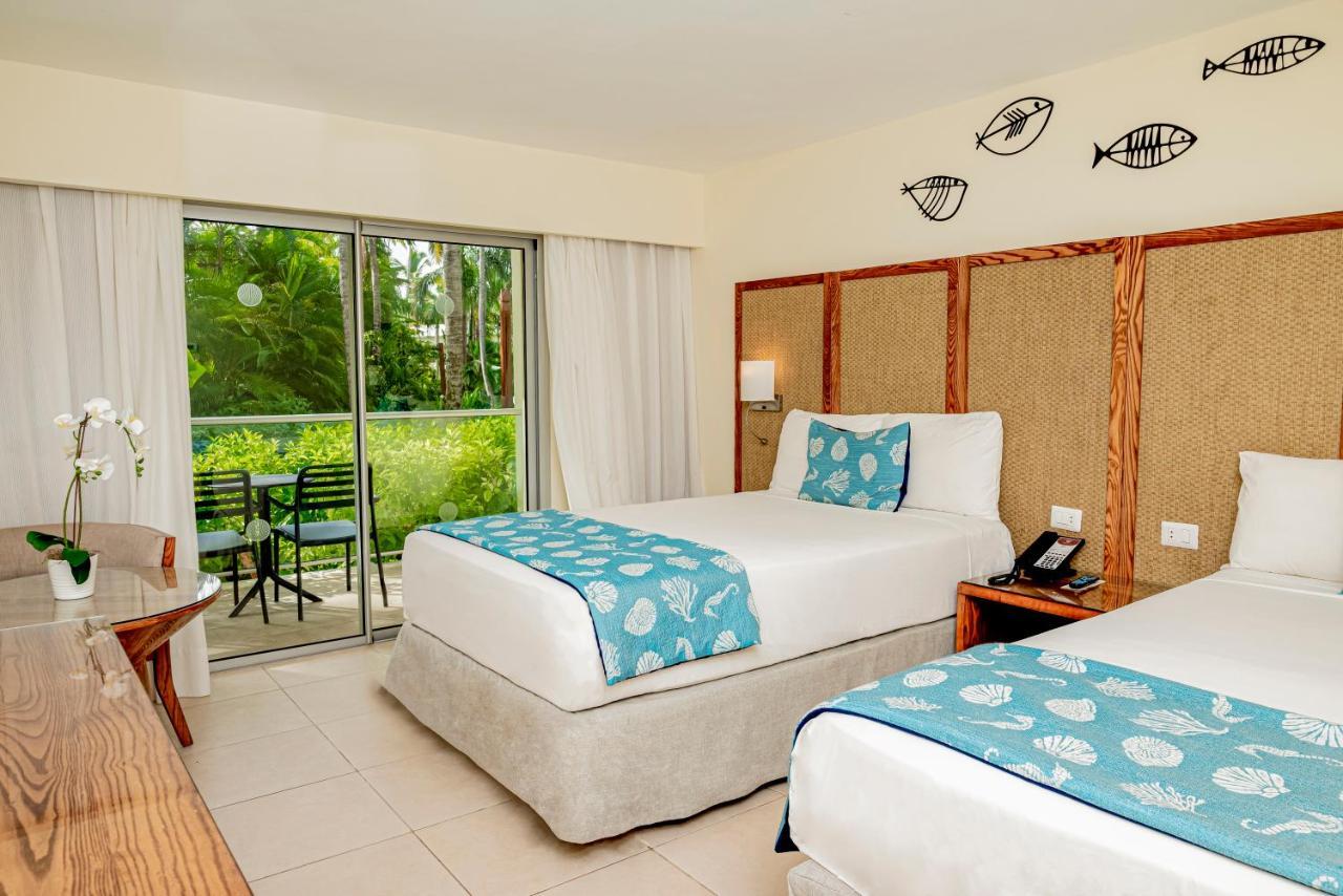 Impressive Punta Cana Hotel Exterior photo The photo depicts a hotel room featuring two beds with white linens and decorative blue throws. The room has a light, neutral color palette and includes wooden accents, such as a headboard and a desk. A large sliding glass door provides access to a b