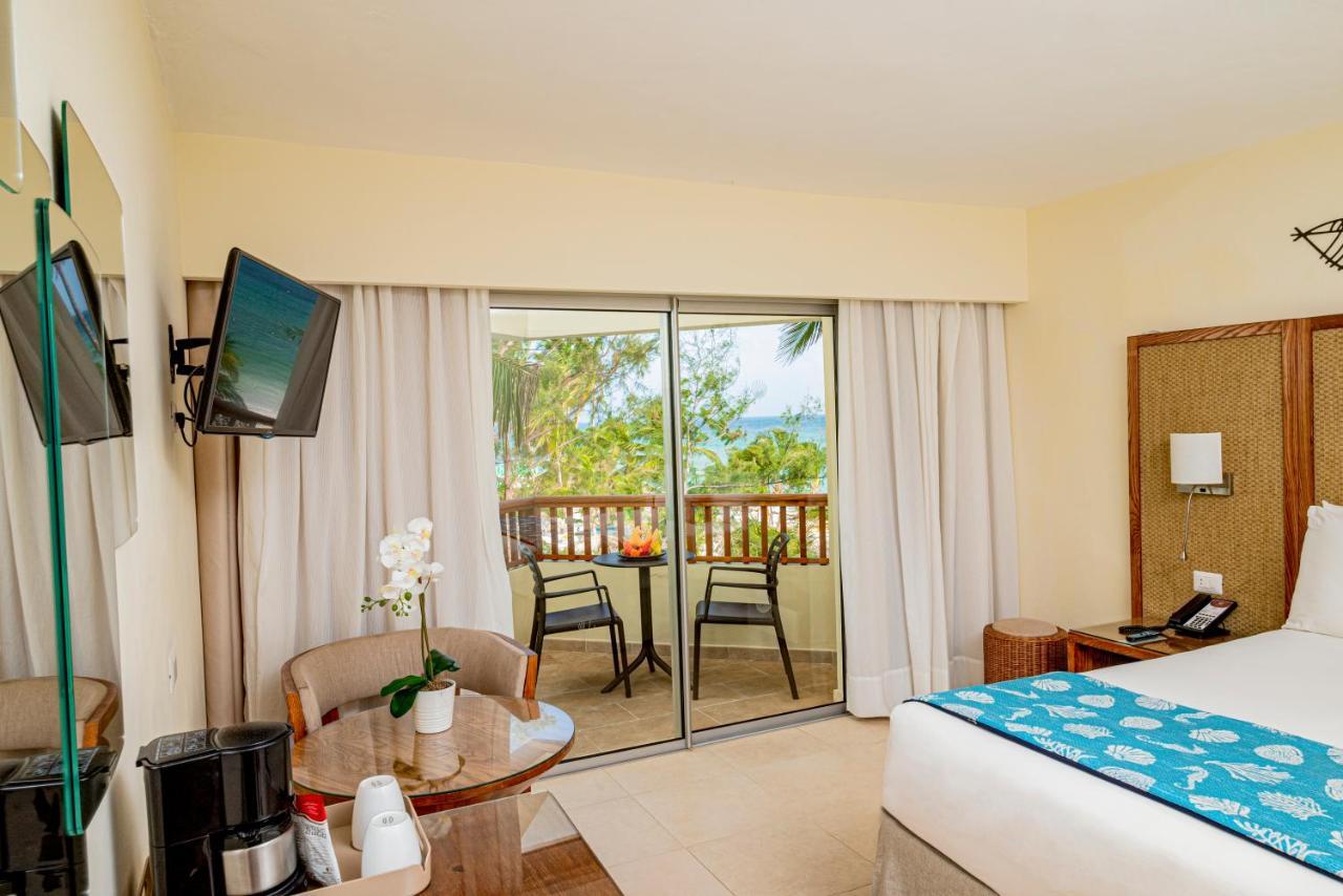 Impressive Punta Cana Hotel Exterior photo The photo shows a hotel room with a light and airy ambiance. There is a bed with a blue and white patterned bedspread. A small round table with a couple of chairs is positioned near a large glass sliding door that leads to a balcony. The balcony offe