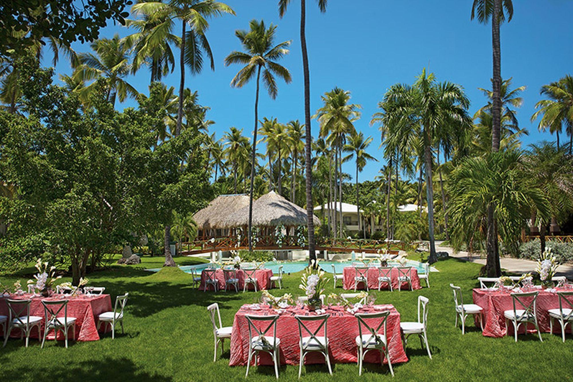 Impressive Punta Cana Hotel Exterior photo The photo depicts a beautiful outdoor setting, likely for an event or celebration. There are several tables arranged on a lush green lawn, each dressed with pink tablecloths. White chairs surround the tables, creating an inviting atmosphere. In the b