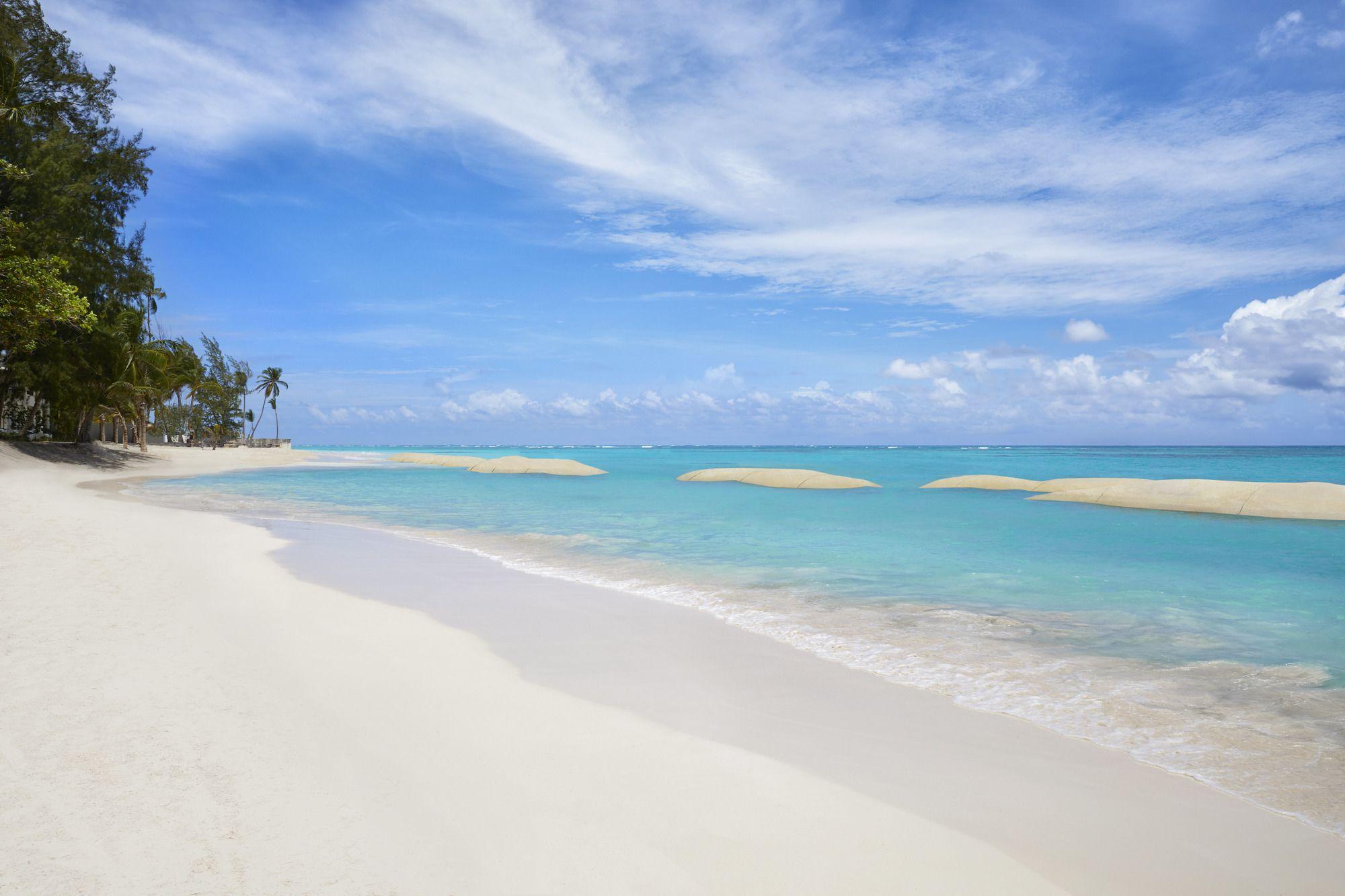 Impressive Punta Cana Hotel Exterior photo The photo showcases a beautiful beach scene. In the foreground, there is soft, white sand leading to clear, turquoise waters. Gentle waves lap at the shore. In the background, palm trees can be seen lining the beach, and the sky is bright with a few 