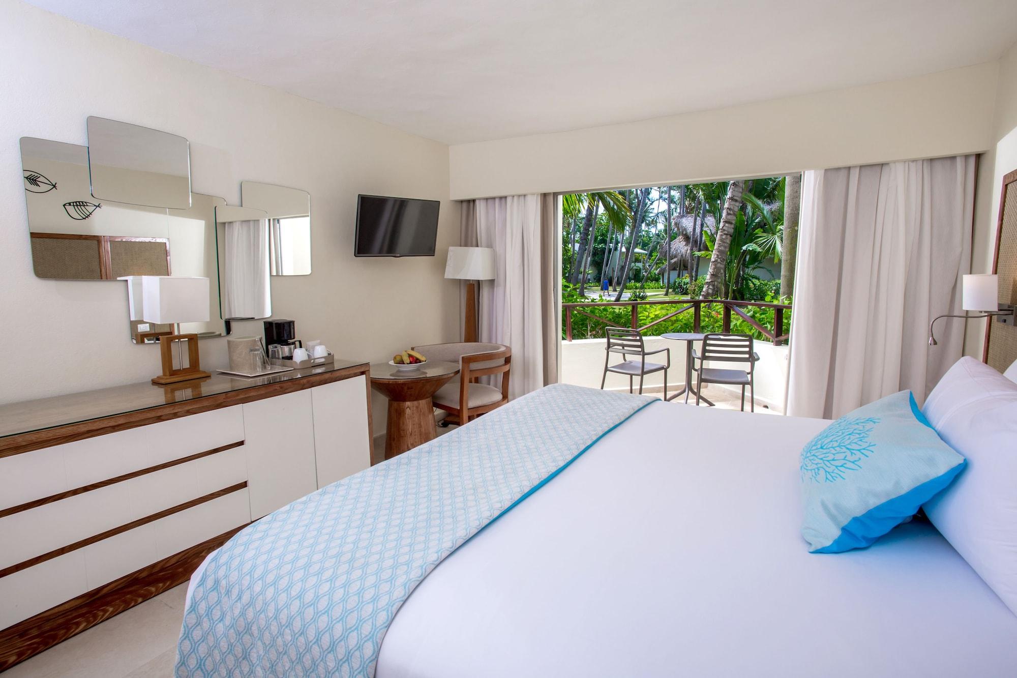 Impressive Punta Cana Hotel Exterior photo The photo shows a well-decorated hotel room featuring a large bed with a light blue throw blanket. There is a bedside table and a dresser with a mirror, as well as a television mounted on the wall. Natural light comes in through a large window that o