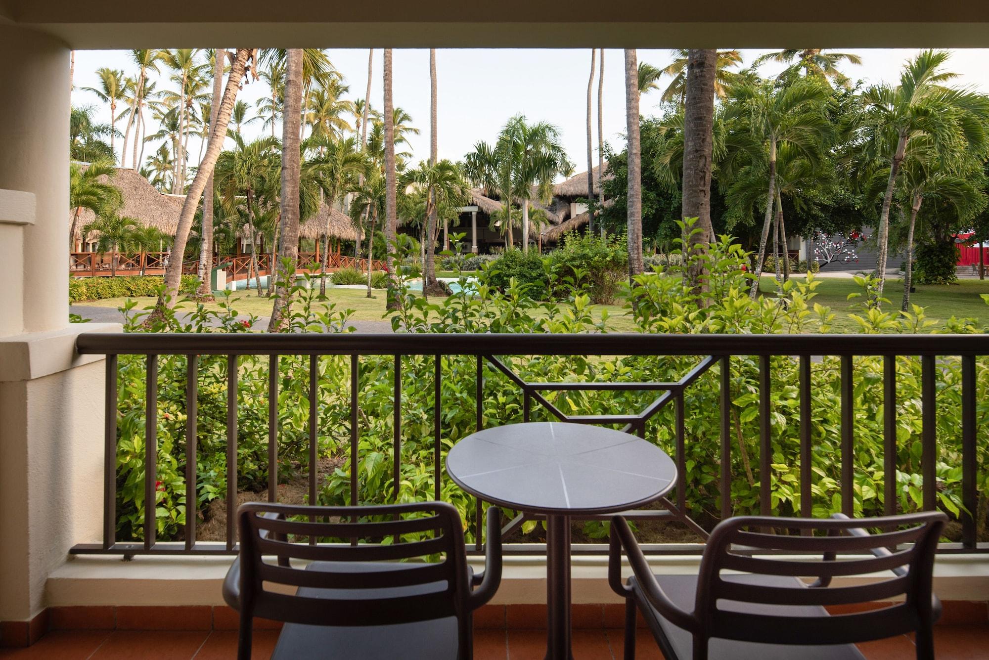 Impressive Punta Cana Hotel Exterior photo The photo shows a view from a balcony with two black chairs and a small round table. In the background, there is a lush green landscape filled with palm trees and other vegetation, suggesting a tropical setting. The area appears well-maintained, with