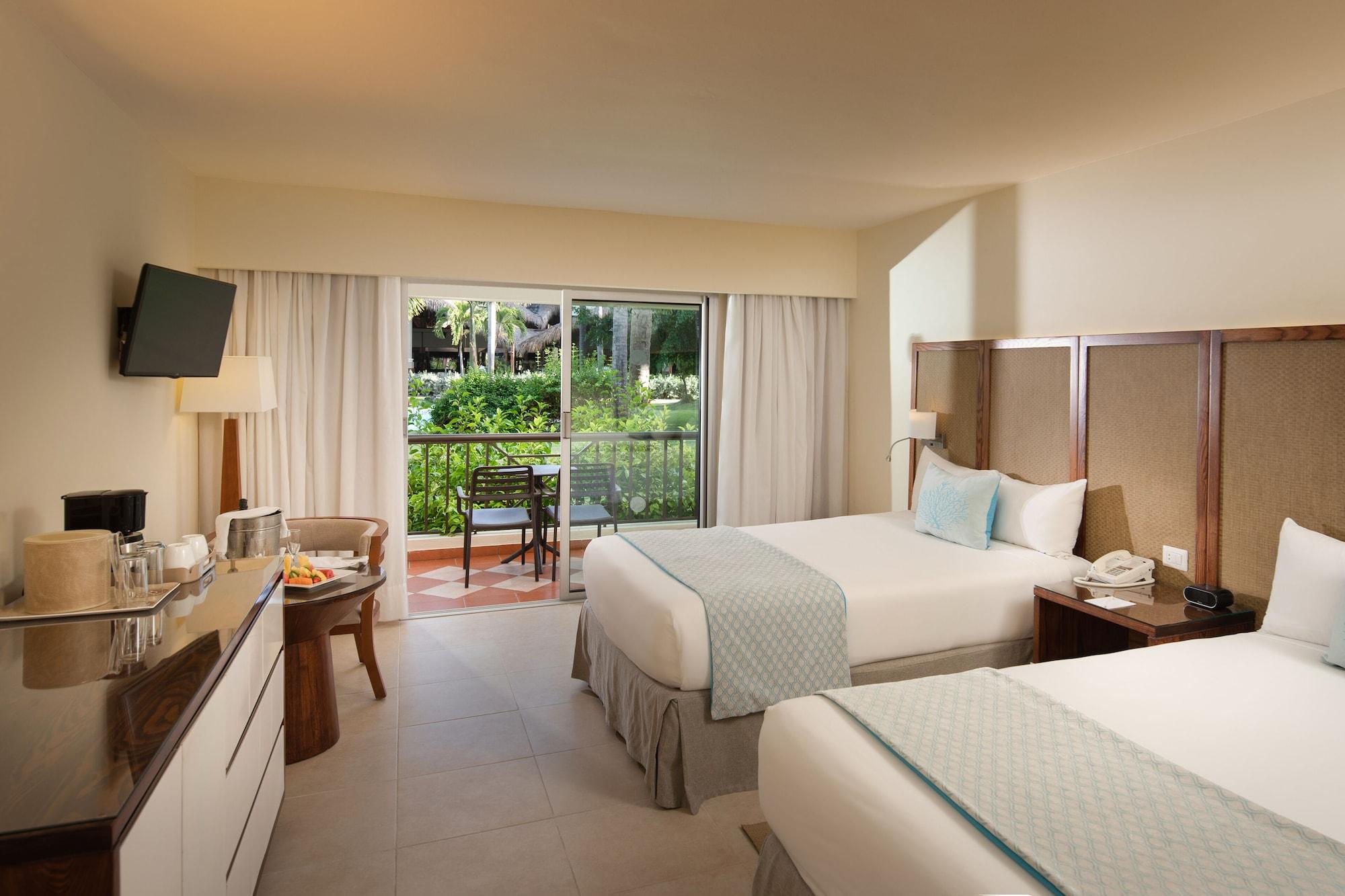 Impressive Punta Cana Hotel Exterior photo The photo shows a hotel room featuring two beds with light-colored linens and decorative pillows. There is a modern armchair beside a small table near a large window that offers a view of an outdoor garden area. A flat-screen TV is mounted on the wal