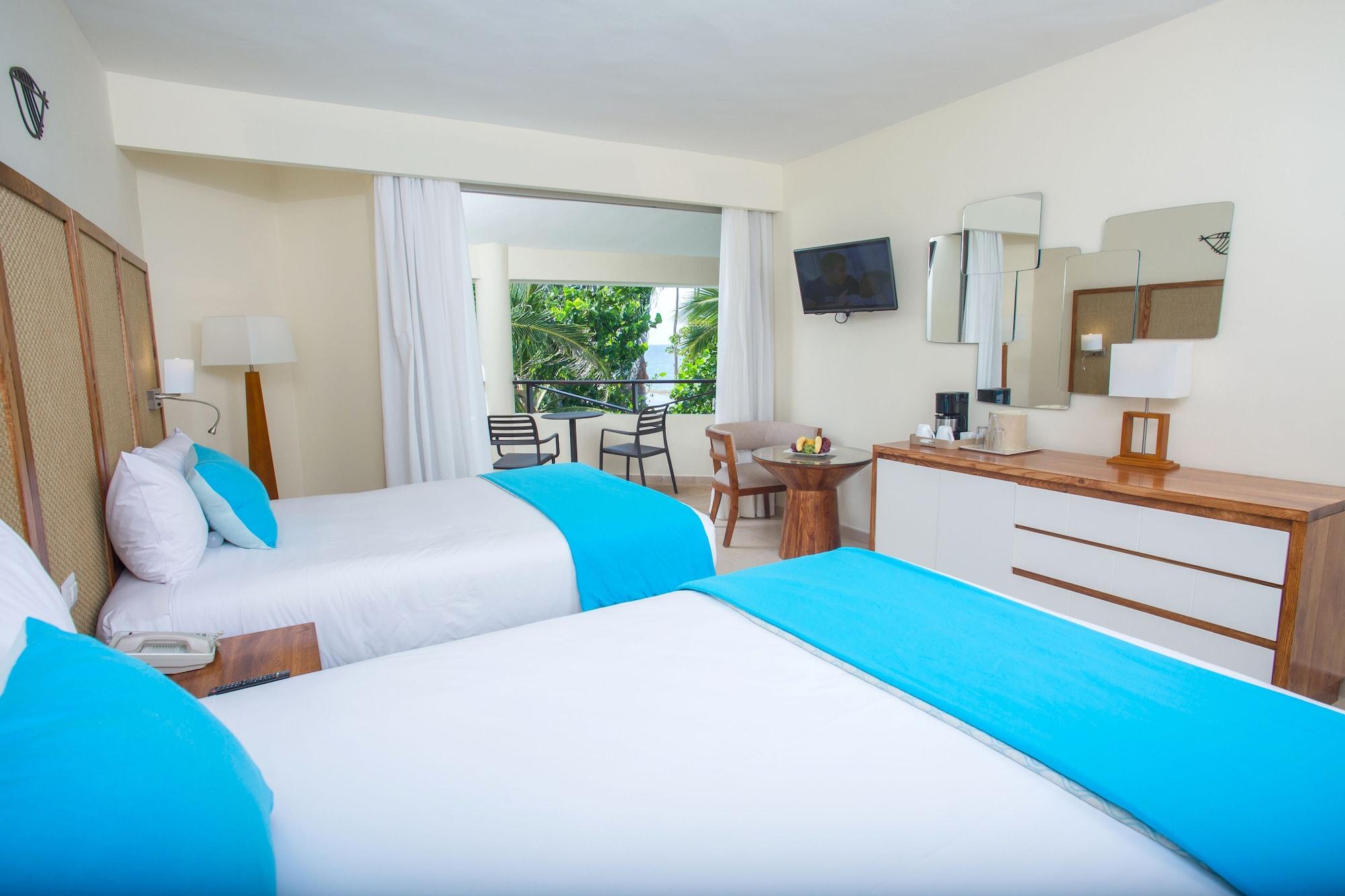 Impressive Punta Cana Hotel Exterior photo The photo shows a cozy hotel room with two beds featuring turquoise bed runners. The room is brightly lit and has light-colored walls. There is a window with sheer curtains that opens to a view of greenery outside. A small table with chairs is positi