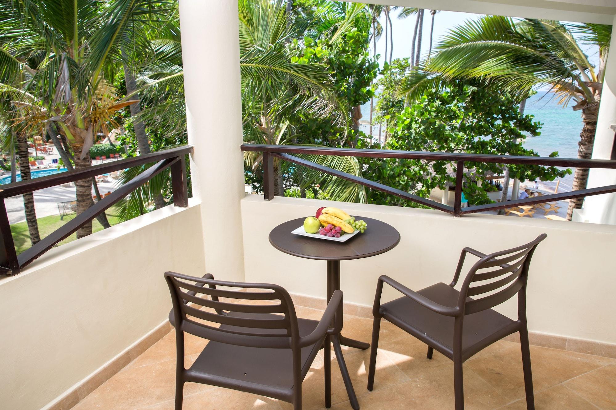 Impressive Punta Cana Hotel Exterior photo The photo depicts a balcony area featuring two black chairs and a small round table. On the table, there is a plate of fresh fruits. In the background, lush green palm trees can be seen, along with a glimpse of blue water, suggesting a tropical or co