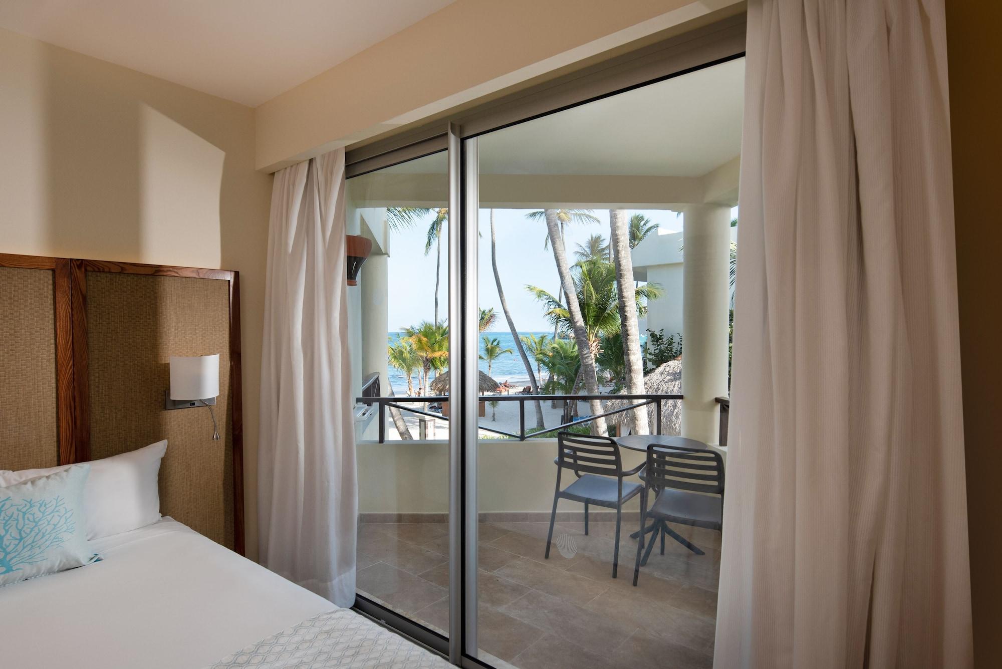 Impressive Punta Cana Hotel Exterior photo The photo shows the interior of a hotel room with a view of an outdoor balcony. On the left, there is a bed with light-colored bedding. A sliding glass door leads to the balcony, which has a couple of chairs and a small table. Outside, you can see pa