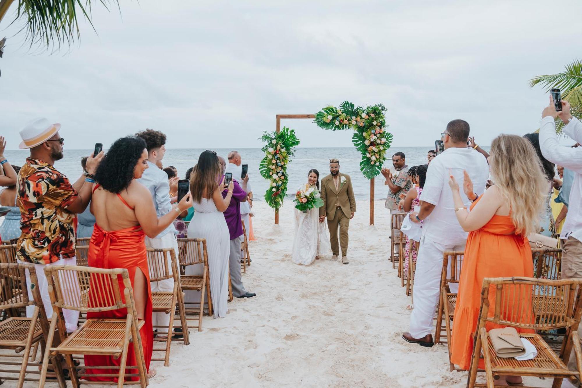 Impressive Punta Cana Hotel Exterior photo The photo depicts a wedding ceremony on a beach. In the center, there is an altar decorated with greenery and flowers. A couple is walking down the aisle, with guests on either side capturing the moment on their phones. The attendees are dressed in a