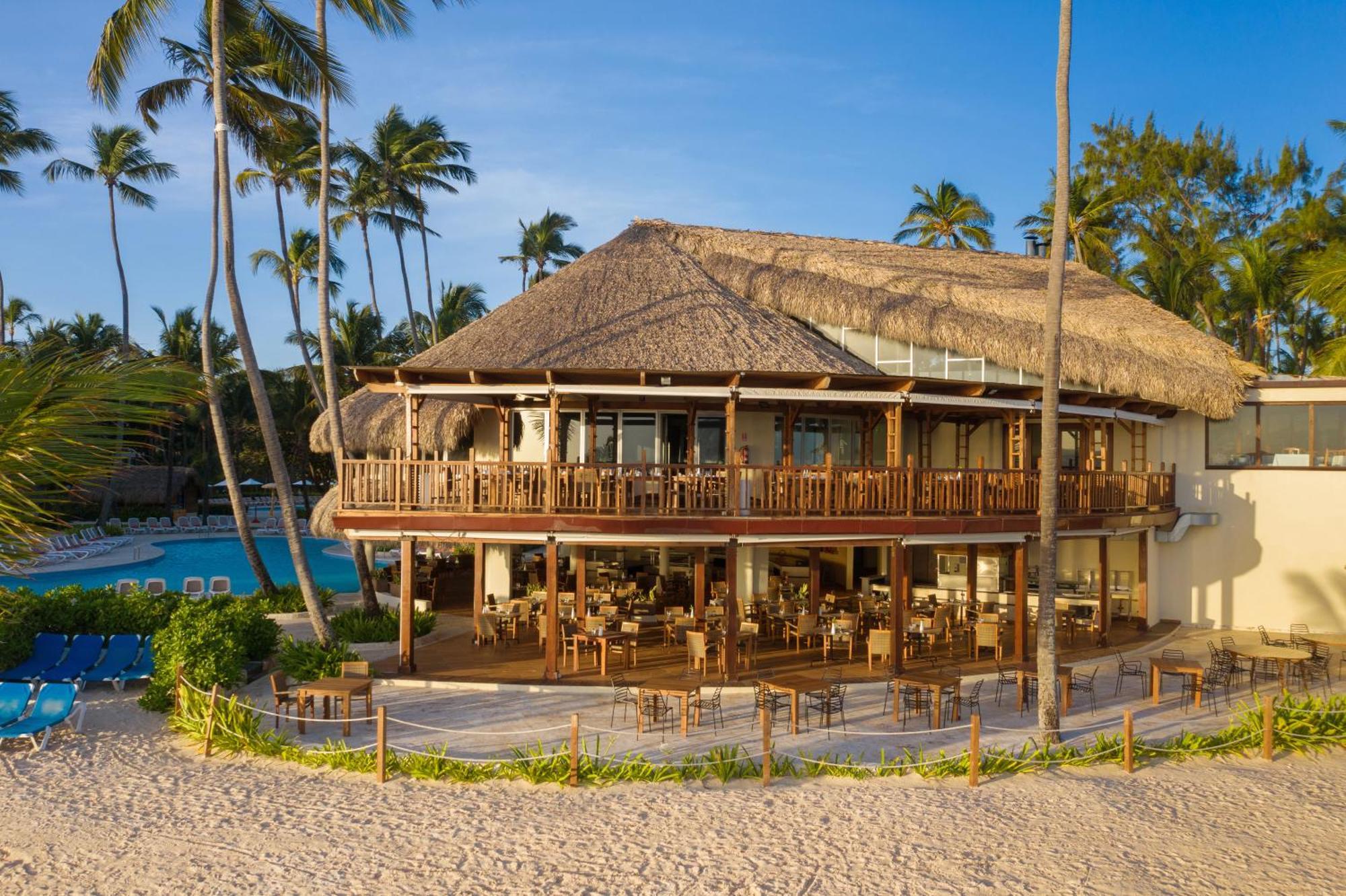 Impressive Punta Cana Hotel Exterior photo The photo shows a tropical beachfront building, likely a restaurant or resort, featuring a large deck with wooden railings and outdoor seating. The structure has a thatched roof and multiple large windows, allowing for a view of the surrounding area.