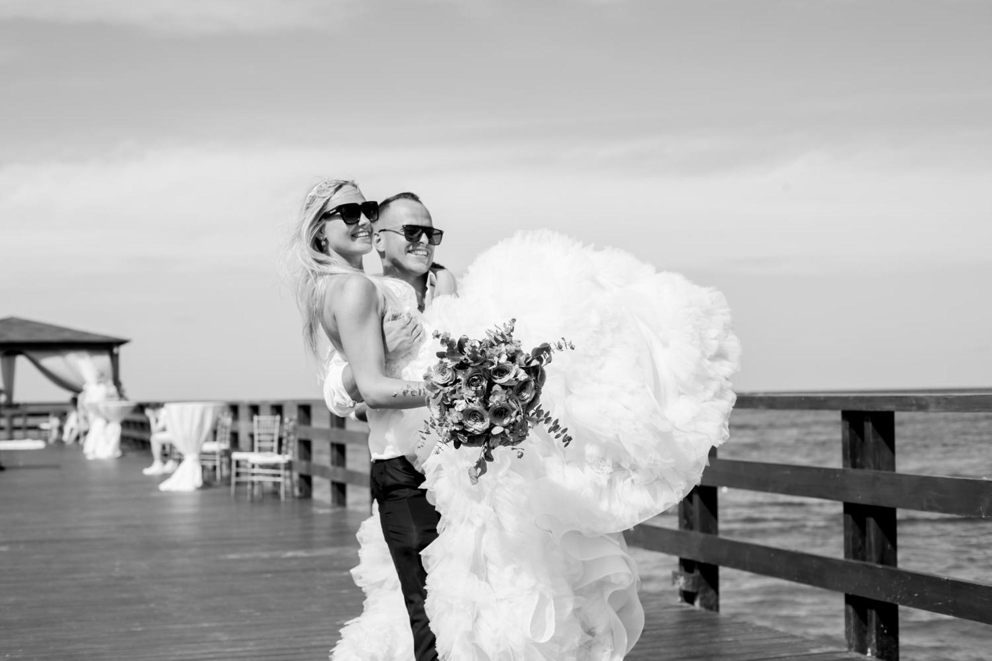 Impressive Punta Cana Hotel Exterior photo The photo appears to depict a couple celebrating their wedding. They are standing on a wooden pier by the water, with the ocean in the background. The scene is in black and white, which adds a classic touch. The bride is wearing a dramatic, fluffy we