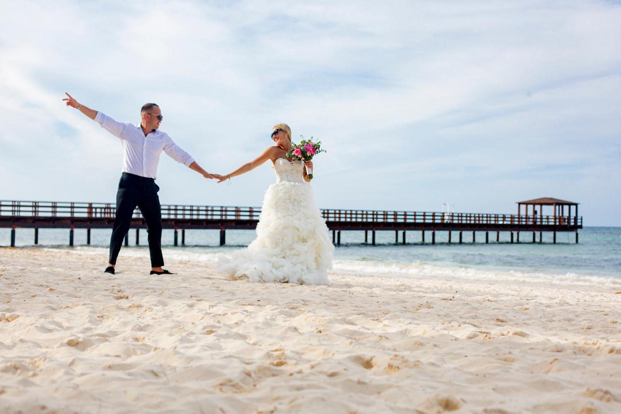 Impressive Punta Cana Hotel Exterior photo The photo depicts a joyful couple on a beach. The bride wears a beautiful white wedding gown and holds a bouquet of flowers, while the groom is dressed in a light shirt and dark pants. They are standing on the sandy shore, holding hands and smiling a