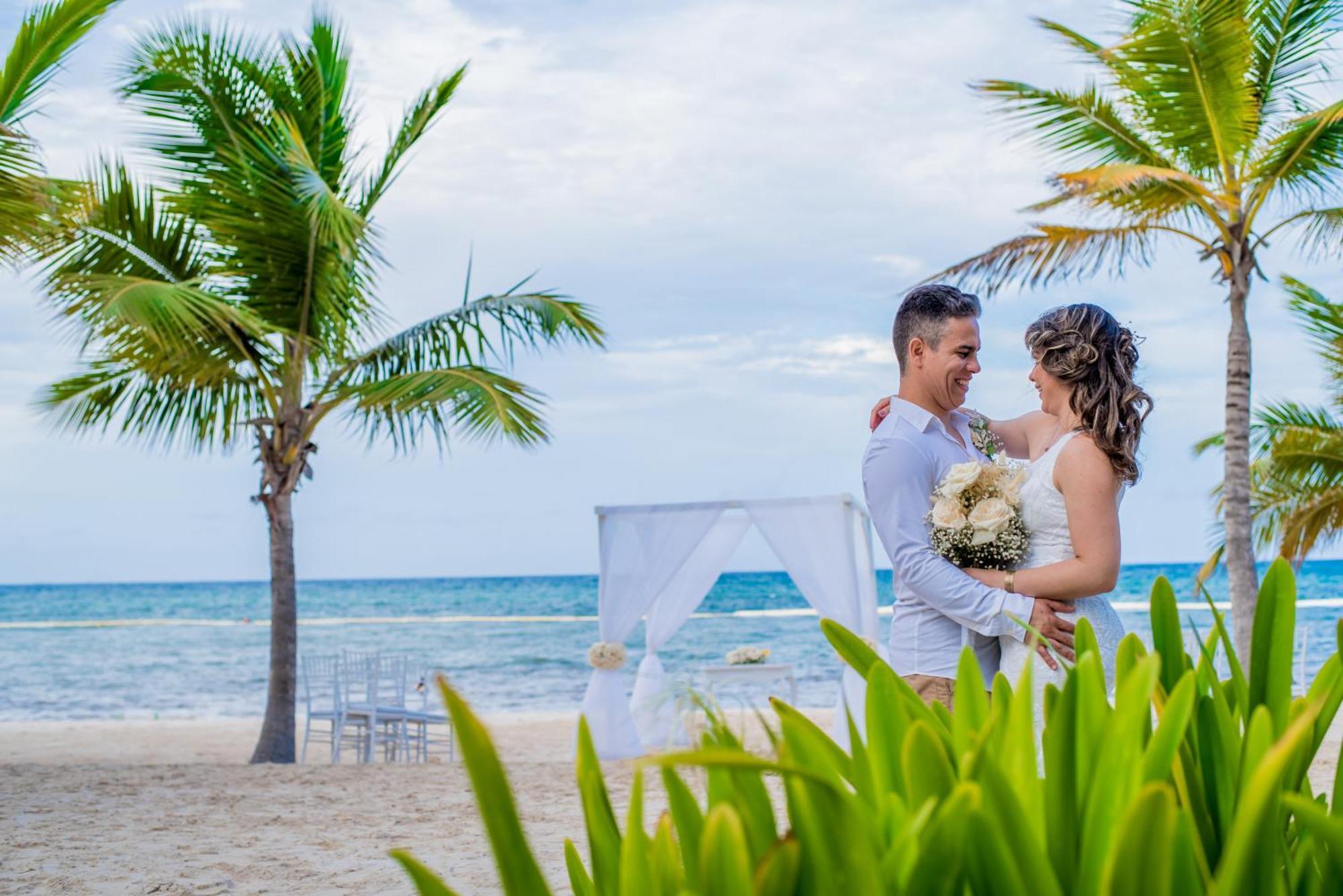 Impressive Punta Cana Hotel Exterior photo The photo depicts a couple embracing on a beach. They appear to be celebrating their wedding, as evidenced by the woman's bouquet. The scenic background features palm trees and the ocean, creating a romantic atmosphere. A white wedding canopy is set 