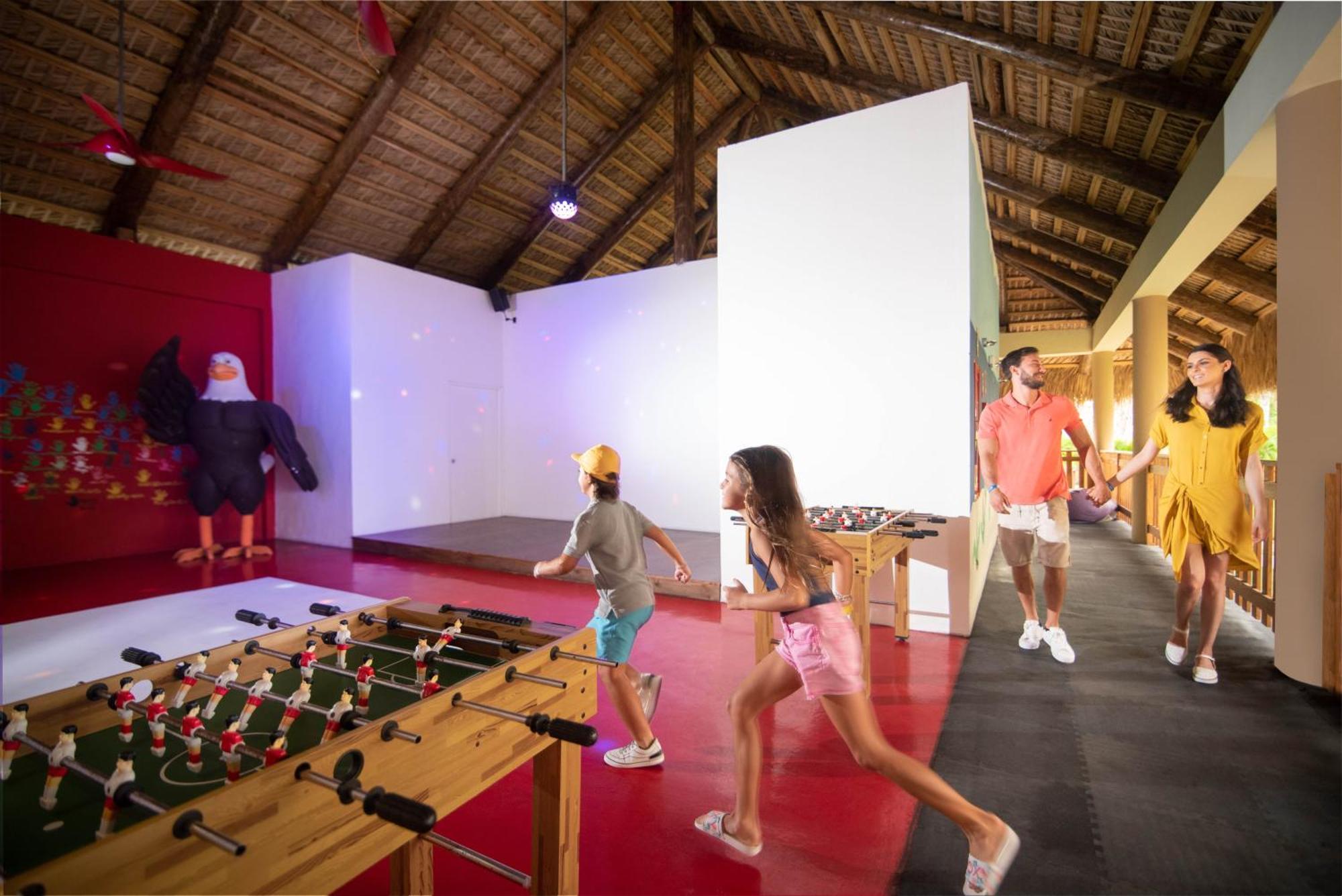 Impressive Punta Cana Hotel Exterior photo The photo shows an indoor recreational space with a thatched roof. In the foreground, there is a foosball table with figures attached to it, where two children are playing or running towards it. A man and a woman, likely parents, are walking together