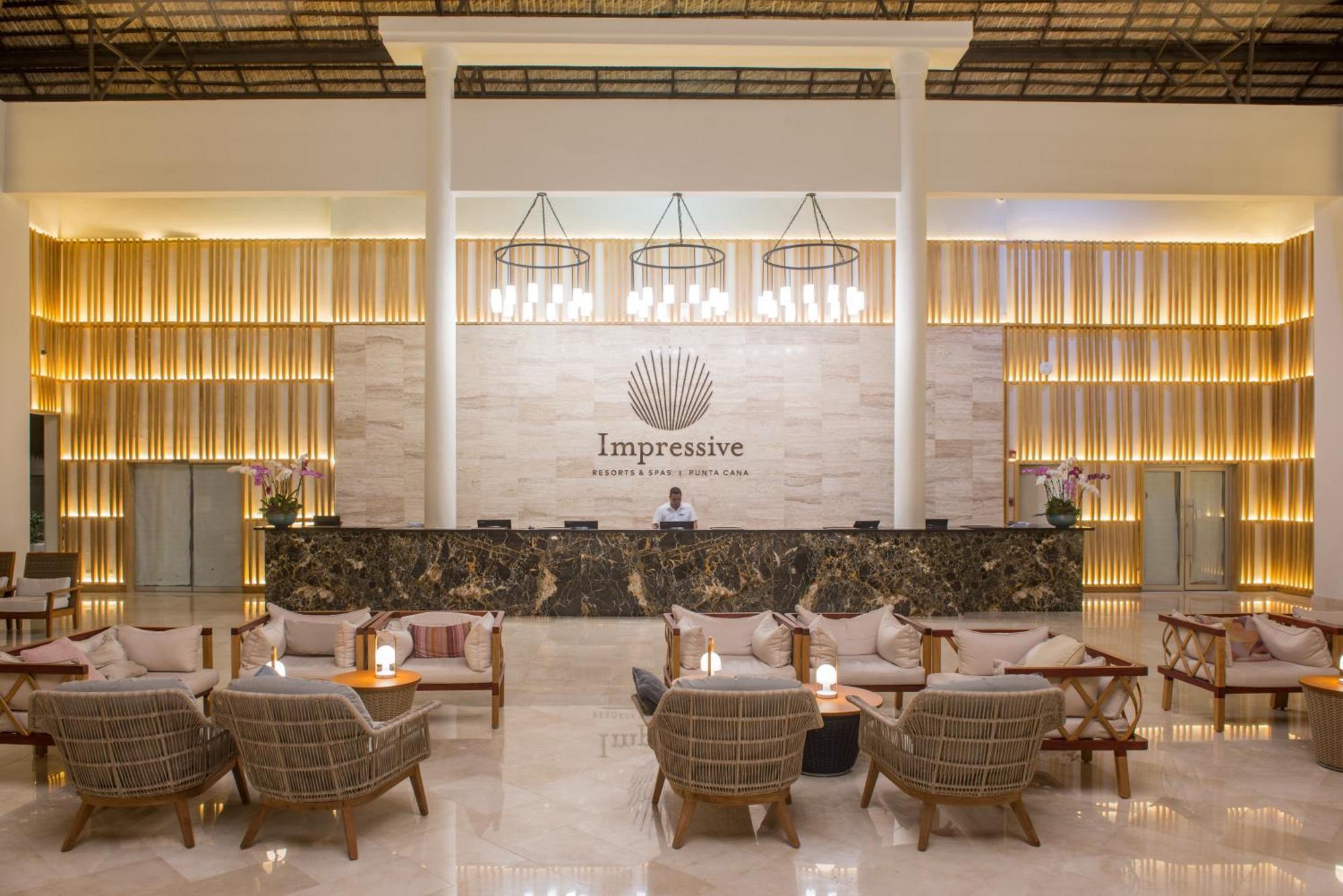 Impressive Punta Cana Hotel Exterior photo The photo shows the interior of a hotel lobby, featuring a modern and elegant design. In the center, there is a reception desk with the word "Impressive" prominently displayed behind it. Above the desk, there are several large light fixtures hanging 