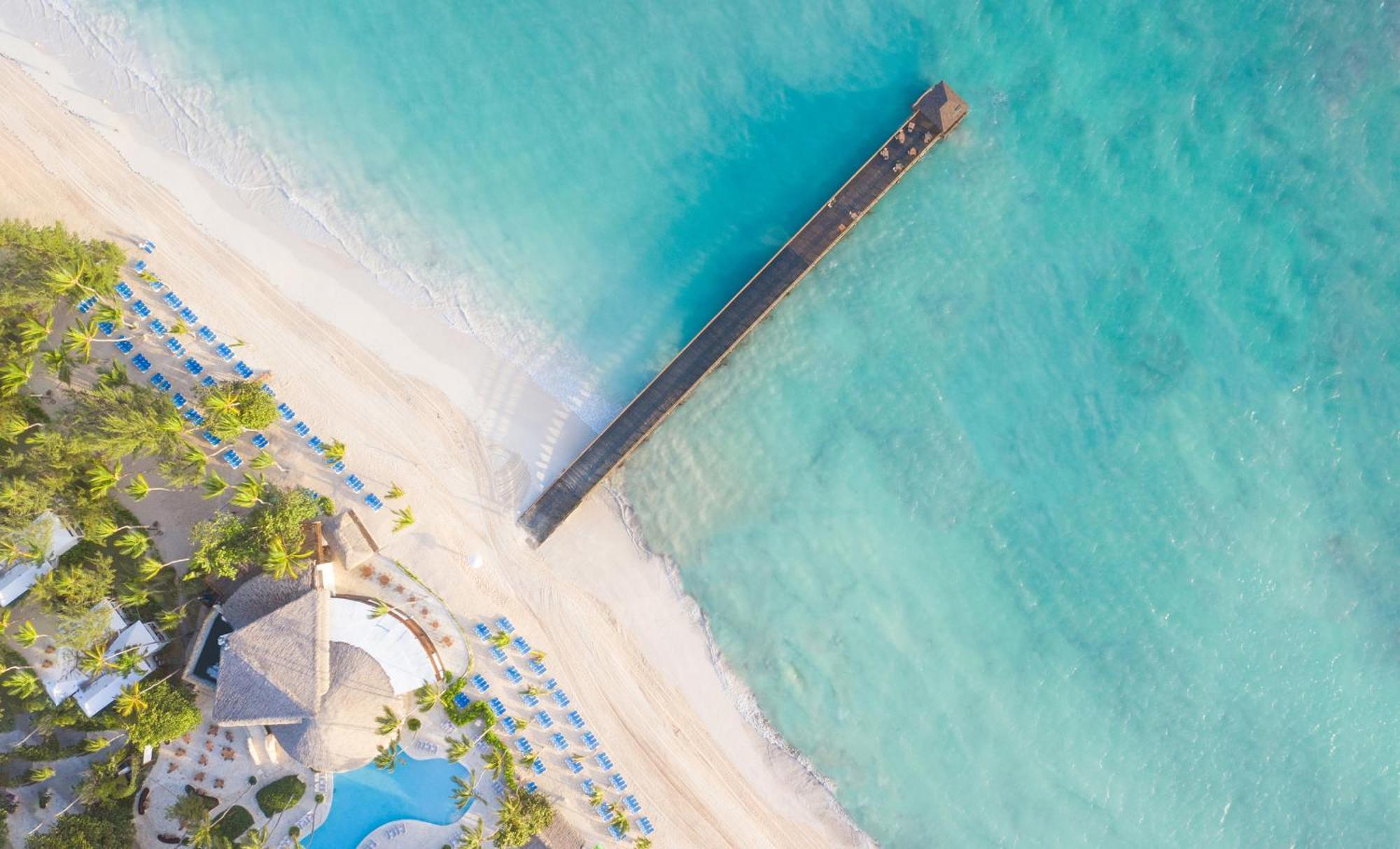 Impressive Punta Cana Hotel Exterior photo The photo shows an aerial view of a beautiful beach scene. There is a long wooden pier extending over clear turquoise waters, leading out from the sandy shore. The beach is lined with umbrellas or lounge chairs, and there is a large building with a t