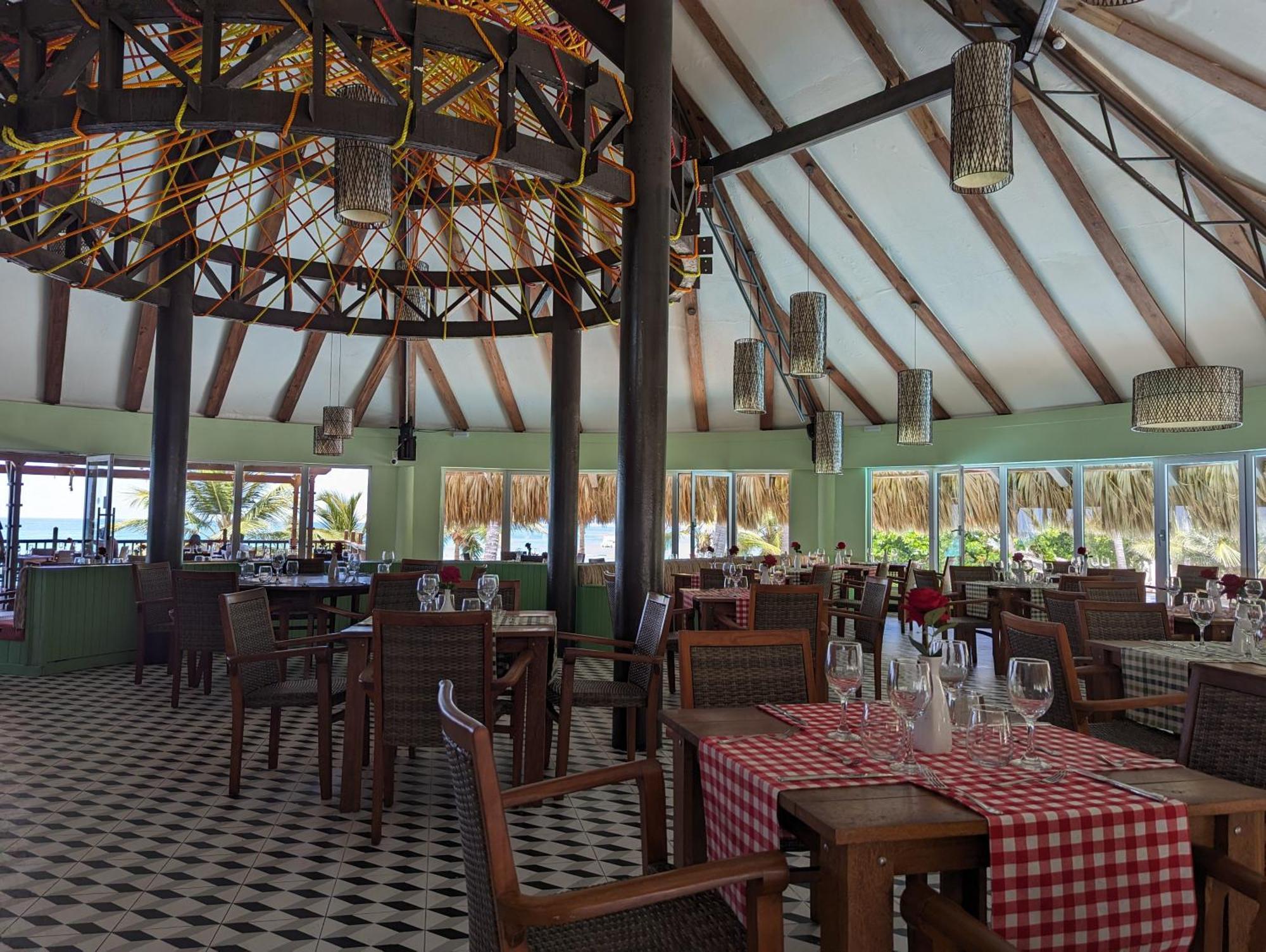 Impressive Punta Cana Hotel Exterior photo The photo shows the interior of a restaurant featuring a tropical or beachy design. The space has high, slanted ceilings with wooden beams and a large decorative light fixture. The floor is tiled in a black-and-white checker pattern. There are wooden