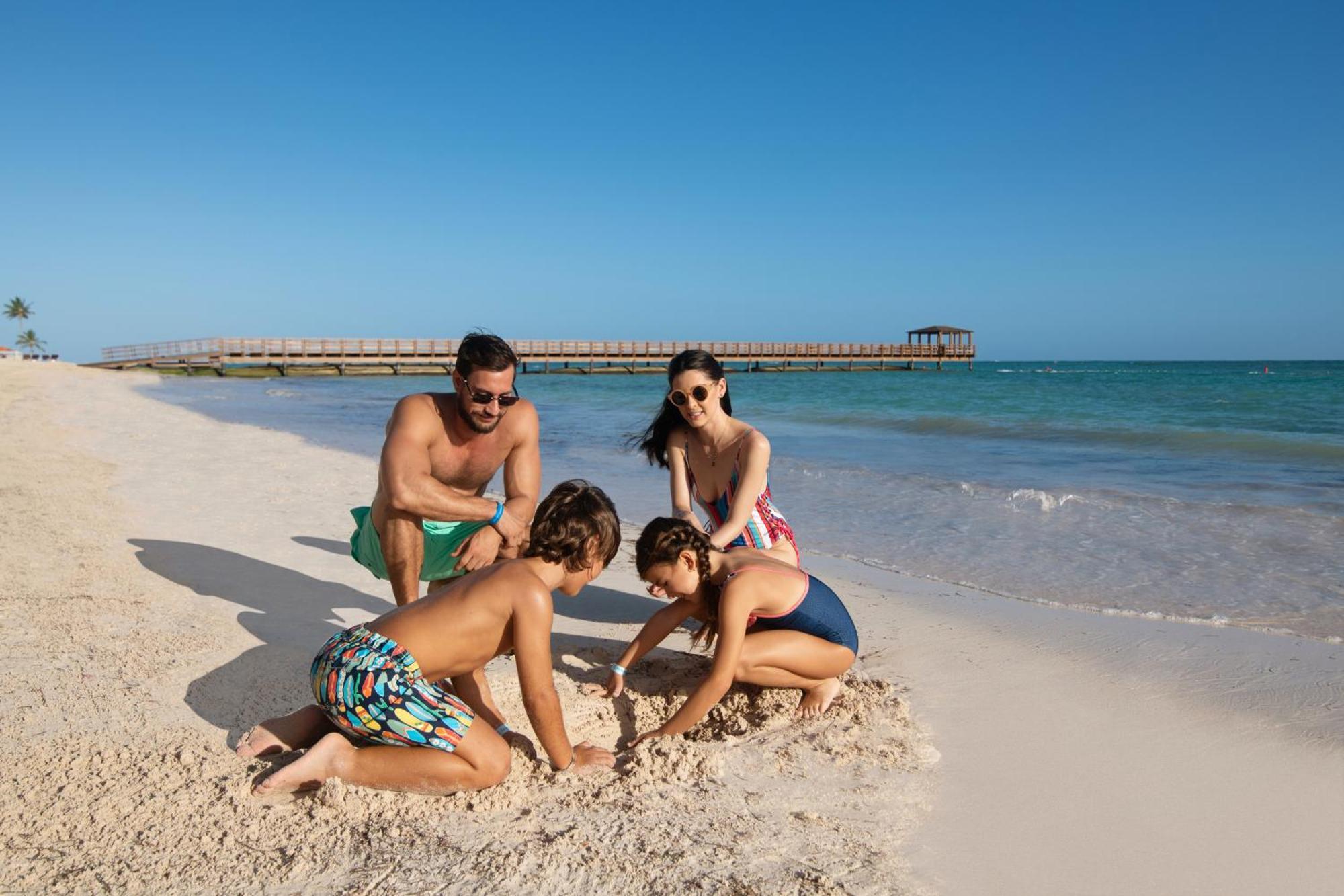 Impressive Punta Cana Hotel Exterior photo The photo depicts a family enjoying a sunny day at the beach. In the foreground, two children are playing in the sand, focused on digging or building something. One child is kneeling, while the other is sitting nearby. An adult male is watching them 