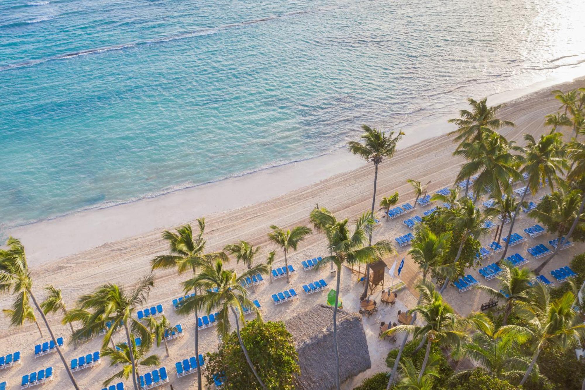 Impressive Punta Cana Hotel Exterior photo The photo shows a picturesque beach scene. There is a wide stretch of sandy beach along the shoreline, bordered by clear, turquoise waters. In the foreground, several palm trees are scattered, adding to the tropical atmosphere. Along the beach, there