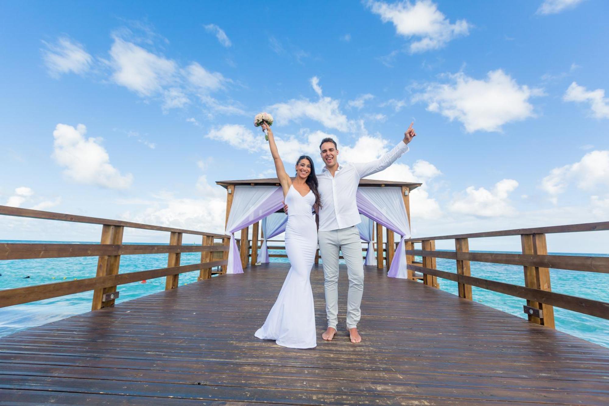 Impressive Punta Cana Hotel Exterior photo The photo shows a joyful couple standing on a wooden pier or deck over the water. The woman is wearing a beautiful white wedding dress and is holding a bouquet of flowers, while the man is dressed in light-colored attire, likely suitable for a beach 