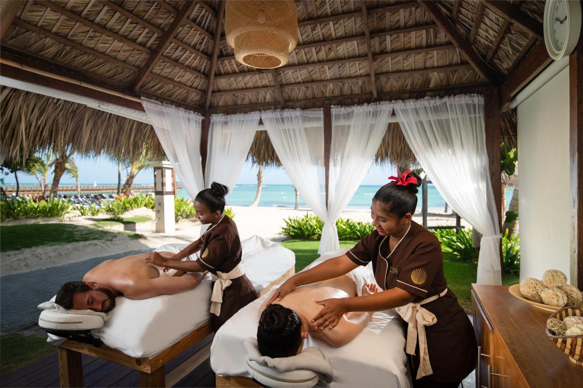 Impressive Punta Cana Hotel Exterior photo The photo depicts a serene outdoor massage setting on a beach. Two therapists are giving massages to clients who are lying on massage tables. The area is shaded by a thatched roof and features sheer white curtains that flutter gently in the breeze. I