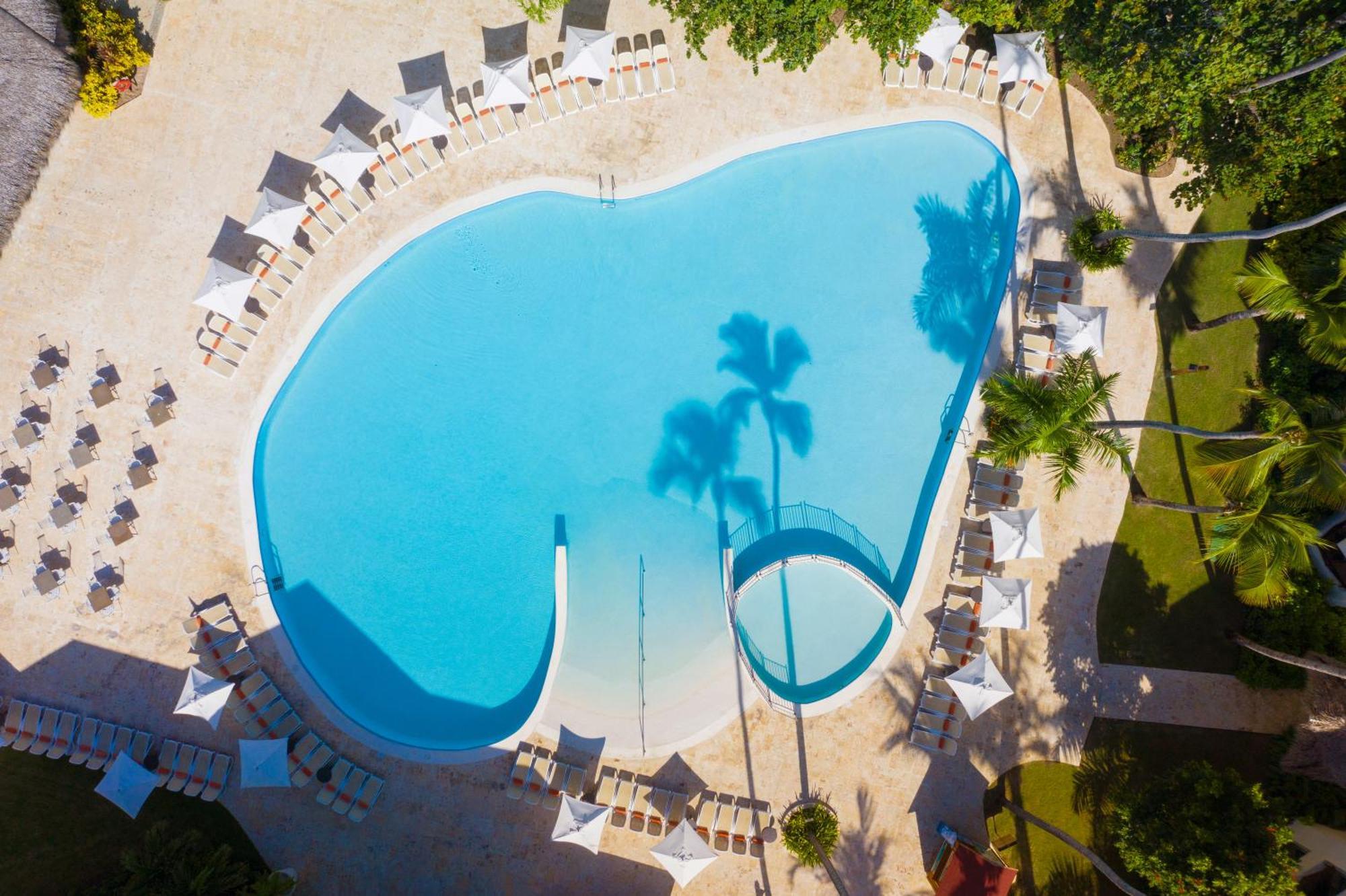 Impressive Punta Cana Hotel Exterior photo The photo shows a large, kidney-shaped swimming pool with clear blue water. Surrounding the pool are several white lounge chairs arranged neatly on a tiled deck. There are palm trees casting shadows on the water, adding to the tropical atmosphere. Th