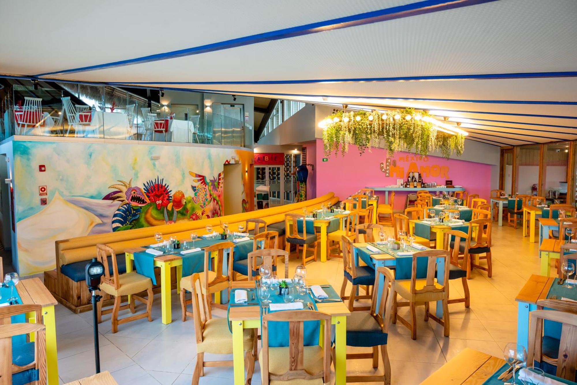 Impressive Punta Cana Hotel Exterior photo The photo shows a vibrant restaurant interior. It features wooden tables and chairs, painted in bright, cheerful colors like yellow and turquoise. The tables are set with utensils and blue tablecloths. The walls are decorated with colorful murals and