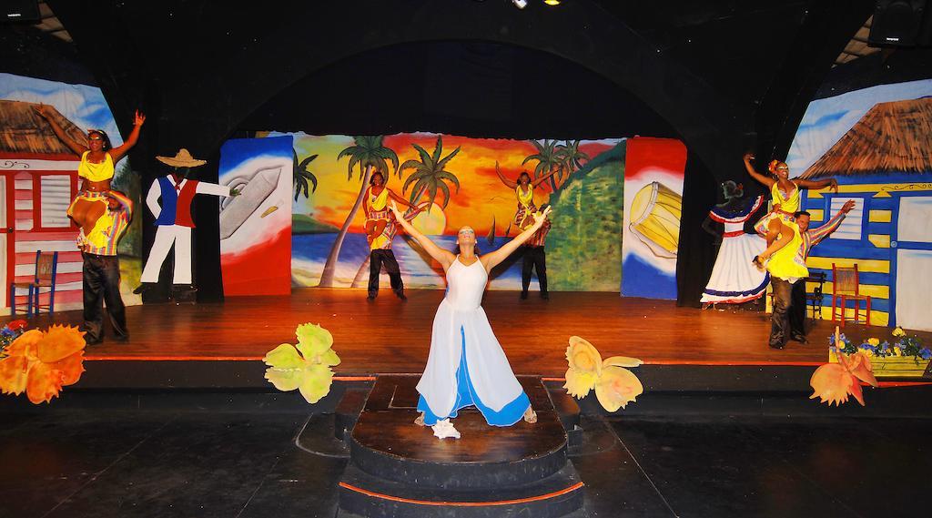 Impressive Punta Cana Hotel Exterior photo The photo shows a vibrant stage performance. In the center, a dancer in a white dress with blue accents is prominently featured, striking a joyful pose. Surrounding her are other performers dressed in colorful costumes, representing various styles of