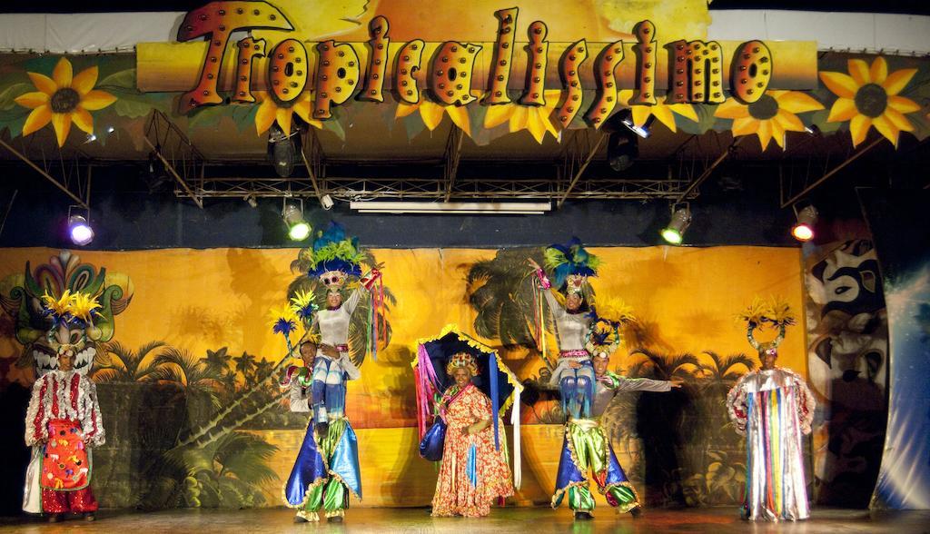 Impressive Punta Cana Hotel Exterior photo The photo shows a colorful stage performance titled "Tropicalissmo." In the image, there are performers dressed in vibrant costumes. Two tall performers on stilts are prominently featured, adorned with elaborate headpieces and outfits that incorporat