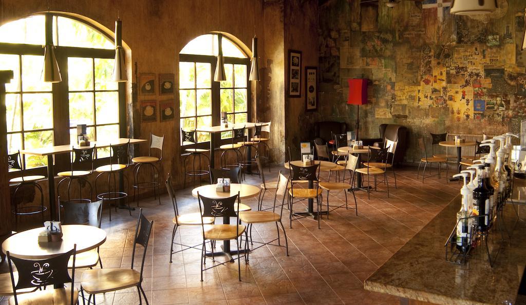 Impressive Punta Cana Hotel Exterior photo The photo shows the interior of a café or restaurant. There are several small tables and chairs arranged throughout the space. Large windows allow natural light to fill the area, creating a warm atmosphere. The walls have a rustic, textured finish wi