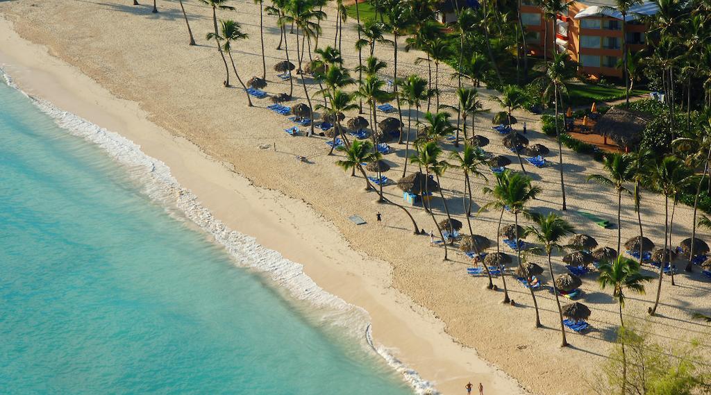 Impressive Punta Cana Hotel Exterior photo The photo shows a picturesque beach scene. In the foreground, there is a sandy shoreline meeting gentle, turquoise waves. Numerous palm trees line the beach, providing shade to sunbathers. There are several beach umbrellas and lounge chairs set up in