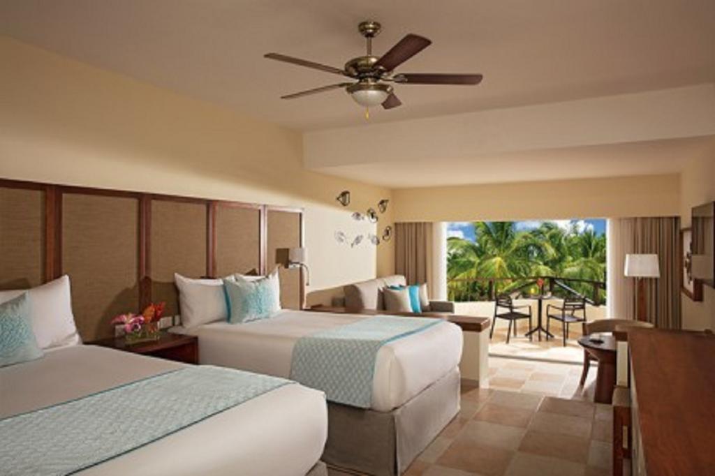 Impressive Punta Cana Hotel Exterior photo The photo shows a hotel room with two beds, decorated in a light, modern style. There is a ceiling fan above and large windows that open to a balcony. The room features soft colors, with a turquoise accent on the bedding, and has a small seating area