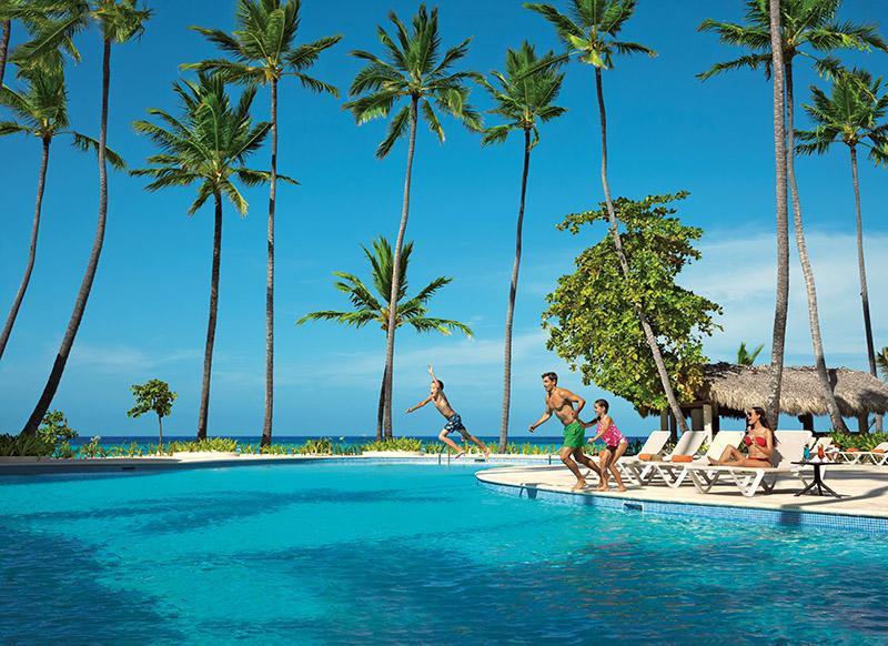 Impressive Punta Cana Hotel Exterior photo The photo showcases a vibrant tropical scene featuring a swimming pool surrounded by tall palm trees. In the foreground, several people are enjoying themselves, with one person jumping into the pool. Others are relaxing on lounge chairs by the poolsi