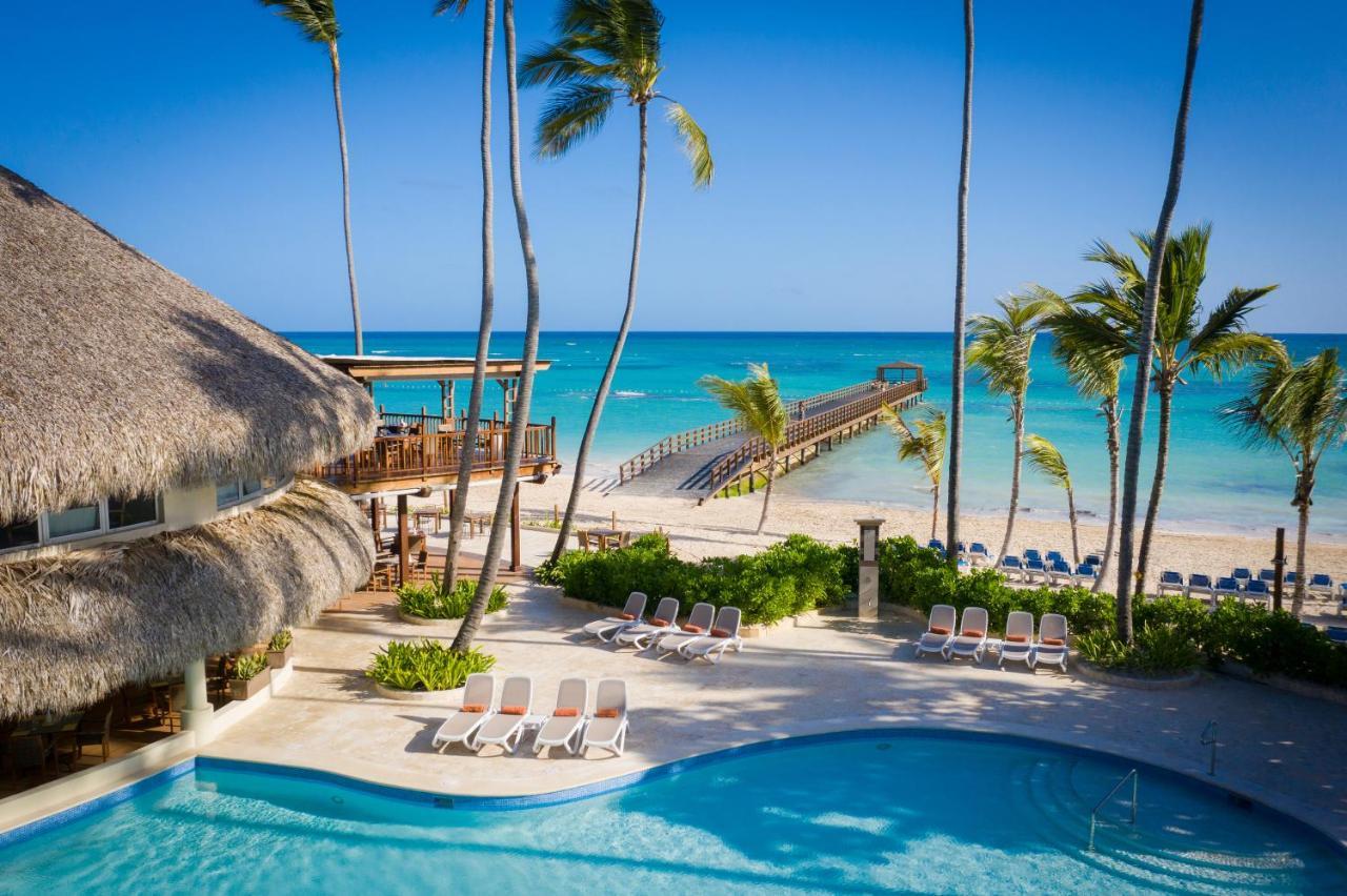 Impressive Punta Cana Hotel Exterior photo The photo depicts a serene tropical scene featuring a swimming pool in the foreground, surrounded by lounge chairs. In the background, there is a beautiful beach with clear turquoise waters and a wooden pier extending out into the ocean. Palm trees f