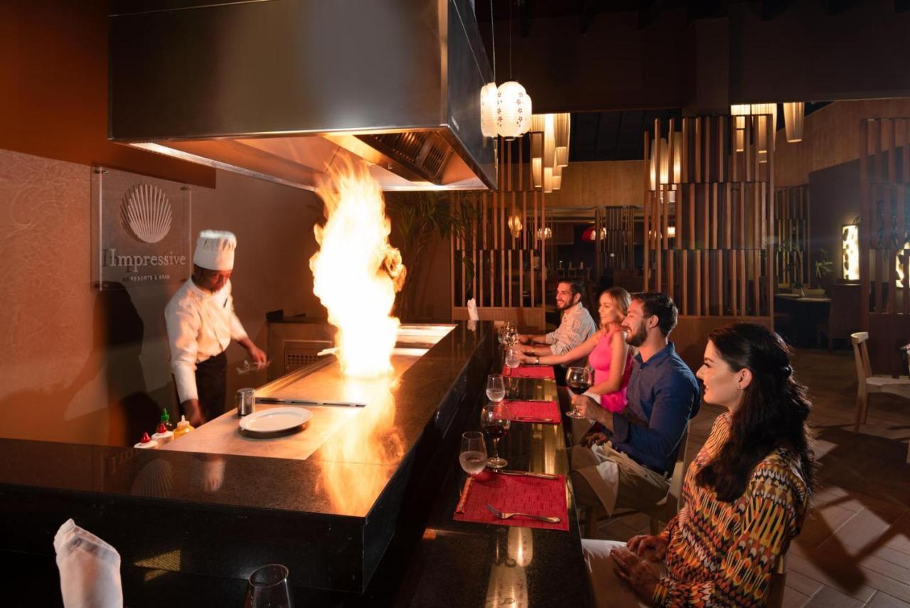 Impressive Punta Cana Hotel Exterior photo The photo shows a dining scene in a restaurant setting. A chef in a white uniform is performing a cooking demonstration at a teppanyaki table, creating a large fire as part of the cooking process. In front of the chef, several diners are sitting at t